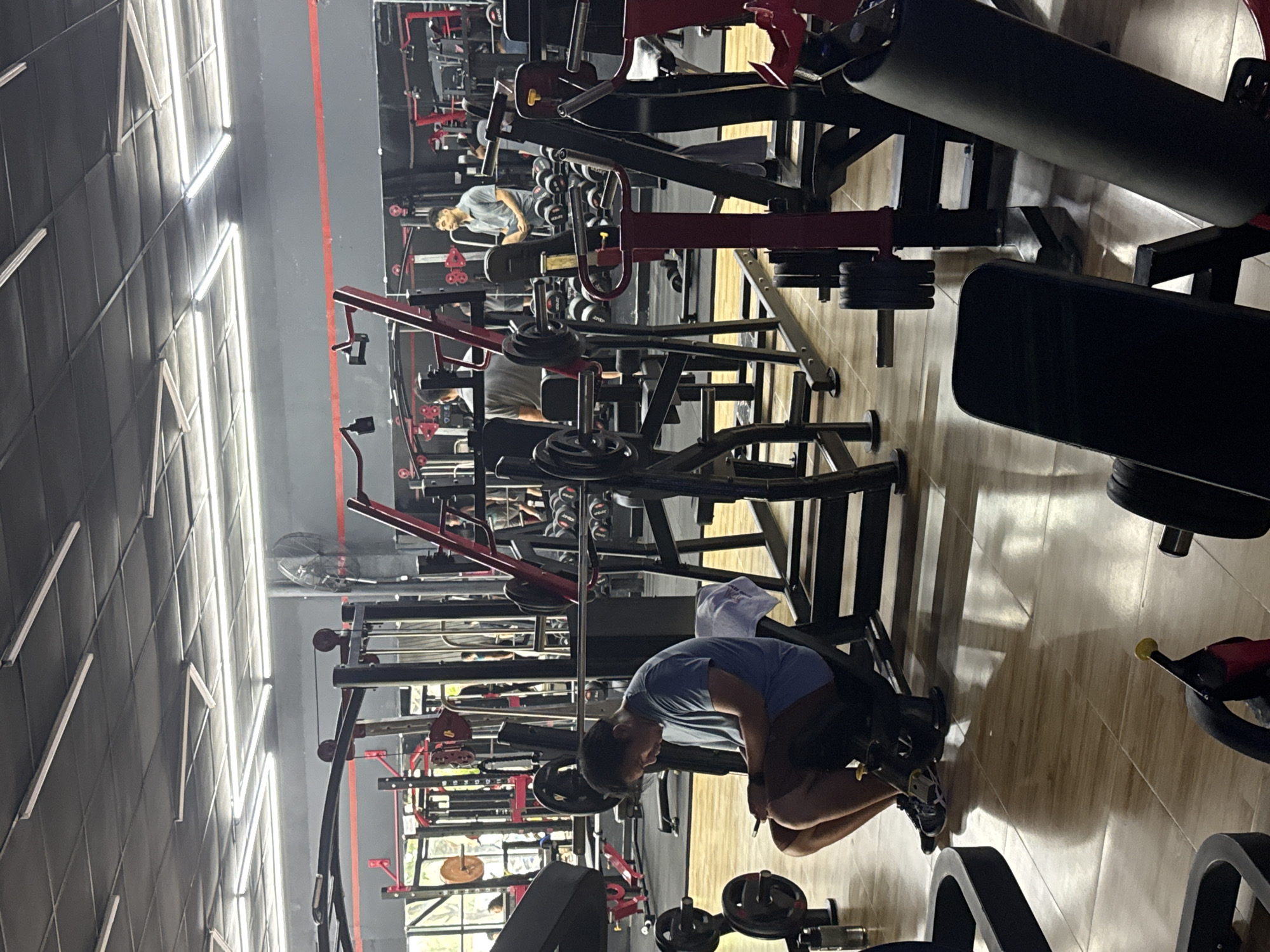 A brightly lit gym in Đà Nẵng, Vietnam features rows of weightlifting machines in black and red. The floor is light-colored tile. A man wearing a blue shirt and shorts is bent over, adjusting or examining something near a weight machine. Other people are seen in the background, using different machines. The gym has a mirrored wall reflecting the equipment and people, and a wall with fluorescent lights.