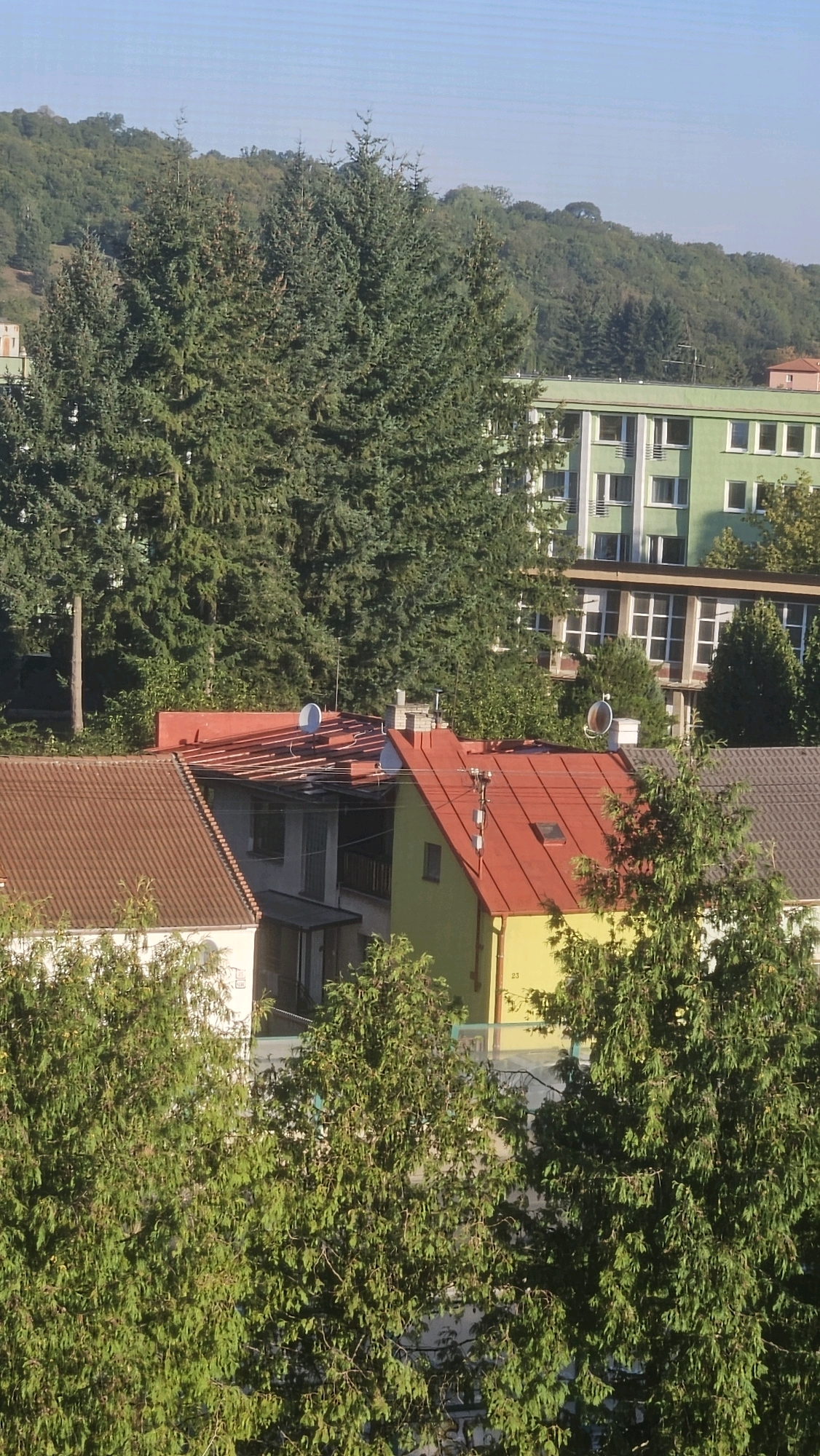This is a high-angle, eye-level medium shot of a residential area in Prešov, Slovakia, on a clear, sunny day. In the foreground, a dense canopy of green trees partially obscures the view of several houses and buildings behind them. The houses have tiled roofs in various colors, including brown, red, and yellow. One house has a bright yellow facade. In the background, a larger, light green building with multiple windows and balconies dominates the scene. Further back, a tree-covered hill rises, and the sky is a clear, pale blue. There are no people visible in the image.