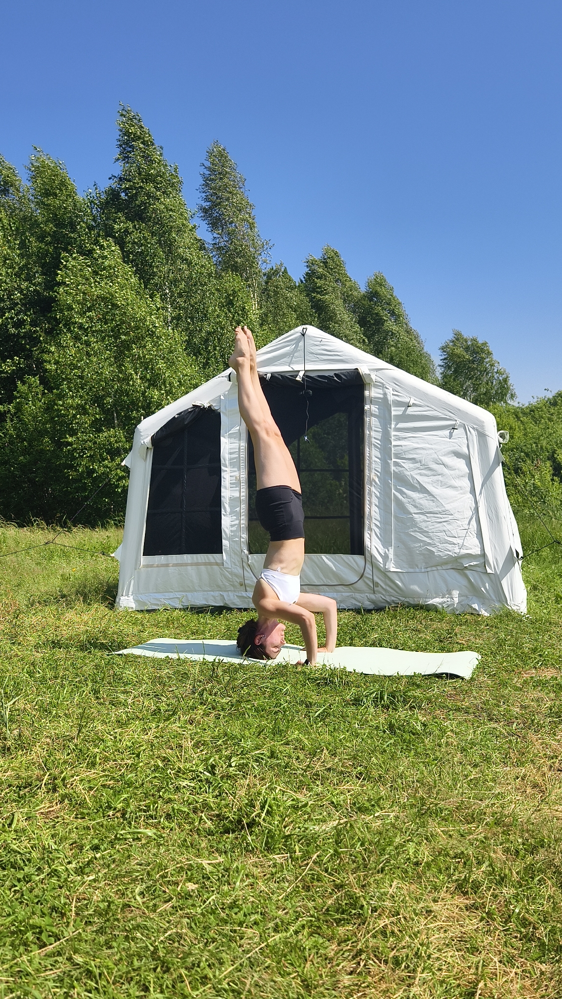 A woman is performing a headstand on a yoga mat in a grassy field. She is wearing a white sports bra and black shorts. Behind her is a large, white tent. The background consists of green trees and a clear blue sky. There is no indication that this scene is in Tyumen, Russia.