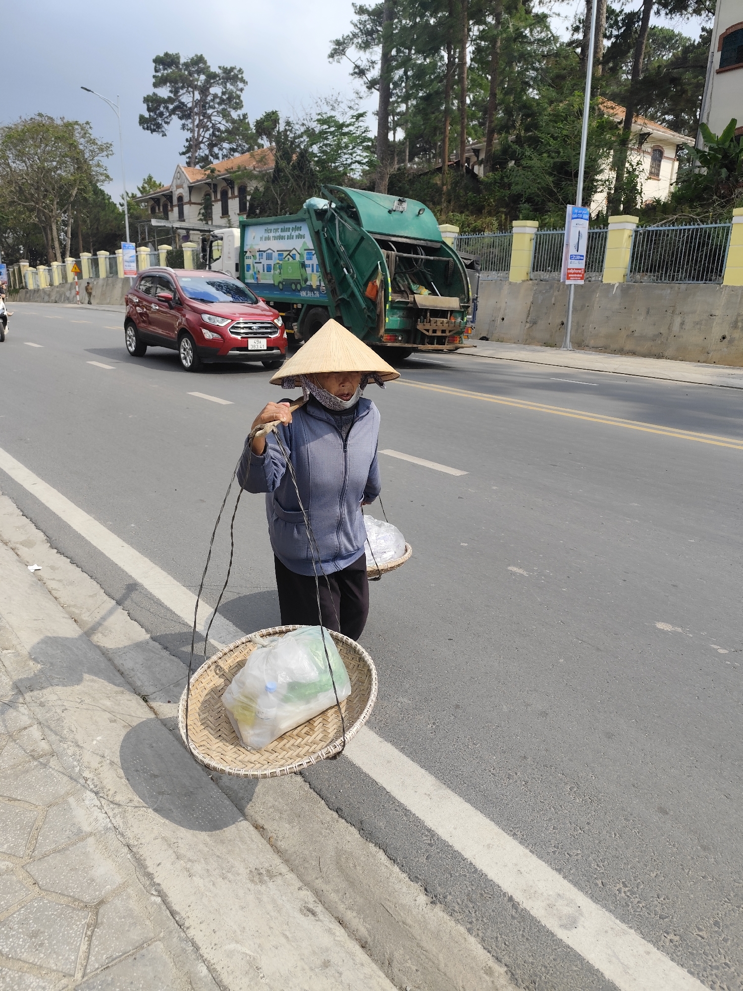 A medium, full outdoor shot captures an individual walking alongside a road in Da Lat, Vietnam. The subject, an older person wearing a traditional conical hat, blue jacket, dark pants, and a face mask, carries two woven baskets suspended from their shoulders by a pole. One basket contains what appears to be refuse in a clear plastic bag. A red SUV drives down the road, along with a green recycling truck. A light gray cement fence runs parallel to the road, behind which are densely spaced trees and buildings. The day appears bright and clear with a slight overcast sky.