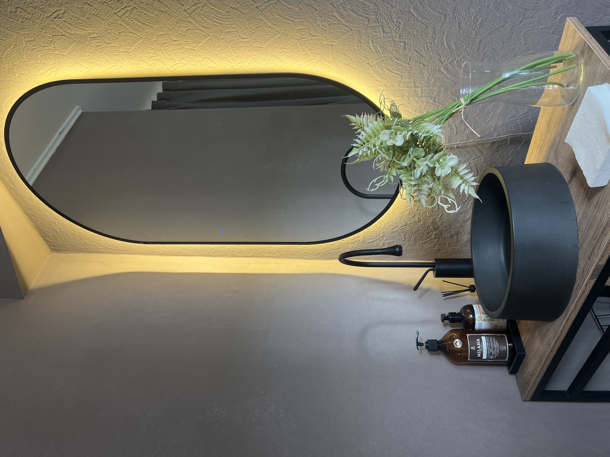 A bathroom scene shows a black oval mirror with an illuminated border hanging on a textured wall. Reflected in the mirror are gray curtains and a portion of the room behind the camera. To the right, a black circular sink is mounted on a wooden vanity with a black metal frame. A bouquet of artificial flowers in a clear glass vase is positioned on the vanity next to the sink. Under the sink, two brown bottles of what appears to be soap or lotion are placed on a shelf. The overall color scheme is muted with tones of gray, brown, and black. No people are visible. The location, Chaikovsky, Russia, is not discernible from the image.