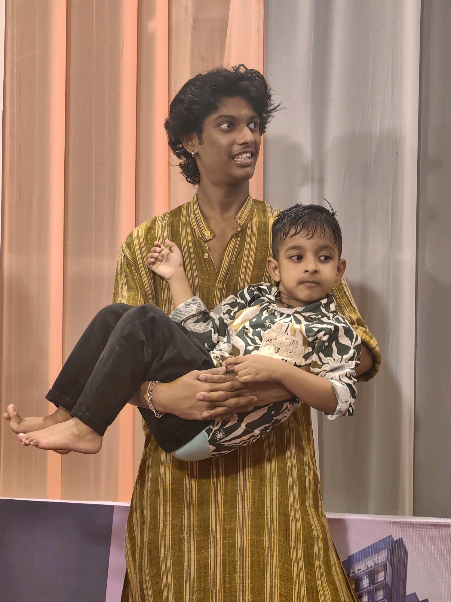 A young man with dark, curly hair, wearing a striped mustard yellow and brown tunic, is holding a younger boy in his arms. The younger boy has dark hair and is wearing a white and green patterned shirt with a beaded necklace. They are standing in front of a backdrop of vertical drapes, one set peach and the other gray. The man's left arm is around the boy's back, and his right arm is supporting the boy's legs. The boy's feet are bare and dangle slightly. Both individuals have a neutral expression. The image appears to be taken indoors, possibly at an event or performance, given the backdrop.