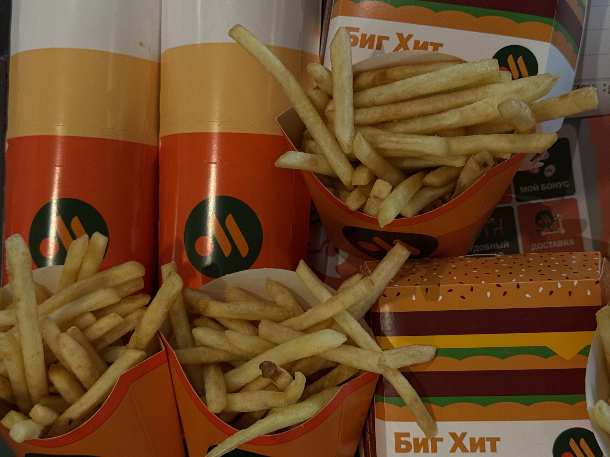 The image shows a close-up of fast food items. There are at least three open cardboard containers filled with french fries. Two cylindrical containers, likely beverages, are visible behind the fries. Boxes with "Биг Хит" (Big Hit) written in Cyrillic are also present. The presence of Cyrillic text and references to "Big Hit" (likely referring to the Big Hit burger at the former McDonald's now named "Вкусно – и точка" in Russia) suggest the scene is likely located in Russia, and based on the text on the box, possibly in the city of Perm. The environment is lit artificially.