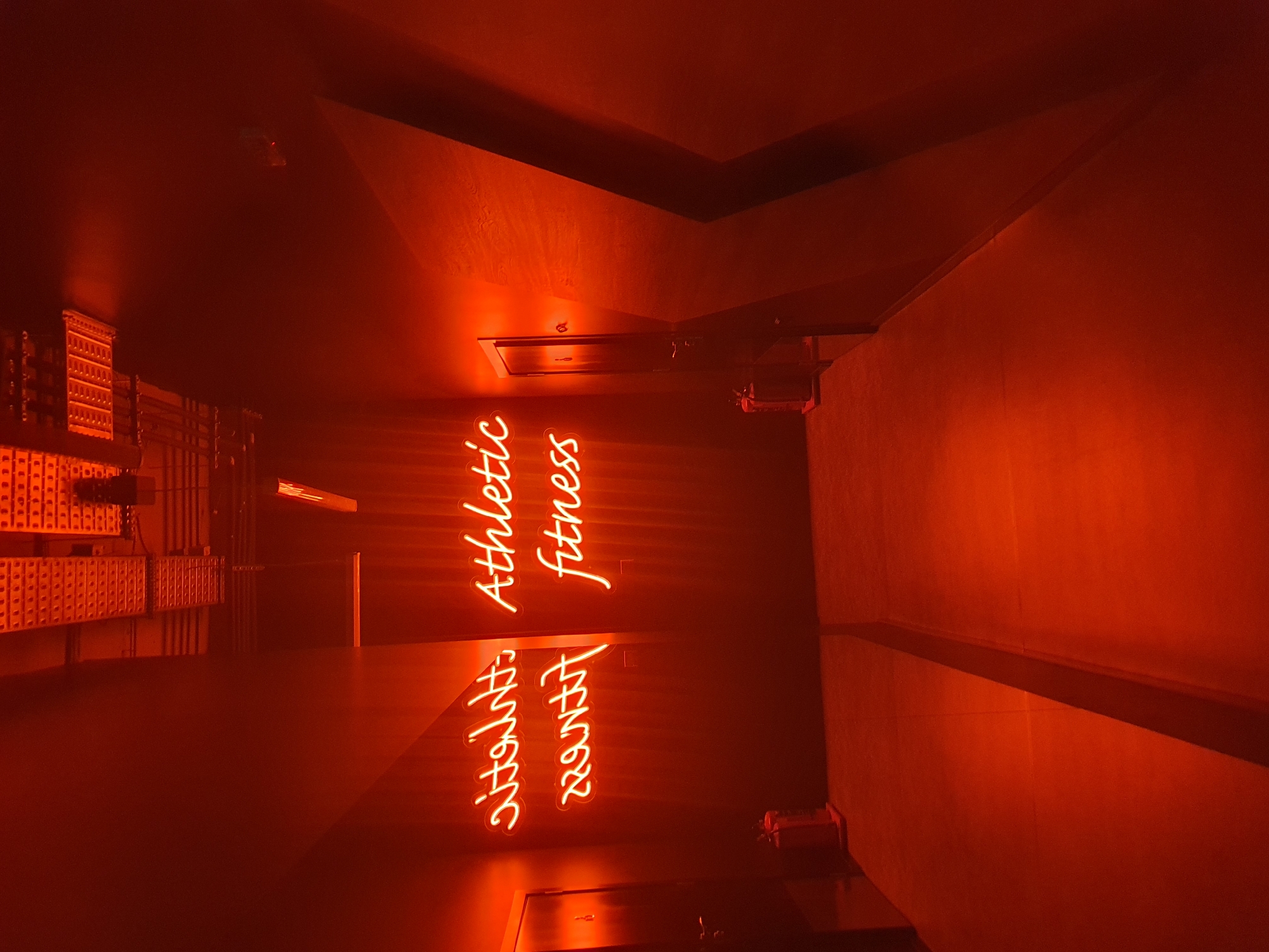 The image depicts the interior of a space with strong red lighting, possibly in Plovdiv, Bulgaria, though this cannot be definitively confirmed from the visual alone.
The phrase "Athletic Fitness" is illuminated in neon lettering, mirrored by its reflection below, along with "Ultimate Workout". The space appears to be a gym or fitness center. Visible are rows of equipment, including what appear to be weights and storage racks. Overhead are vents and some lighting fixtures. The overall scene is dark, dominated by the red light, making specific details challenging to discern.