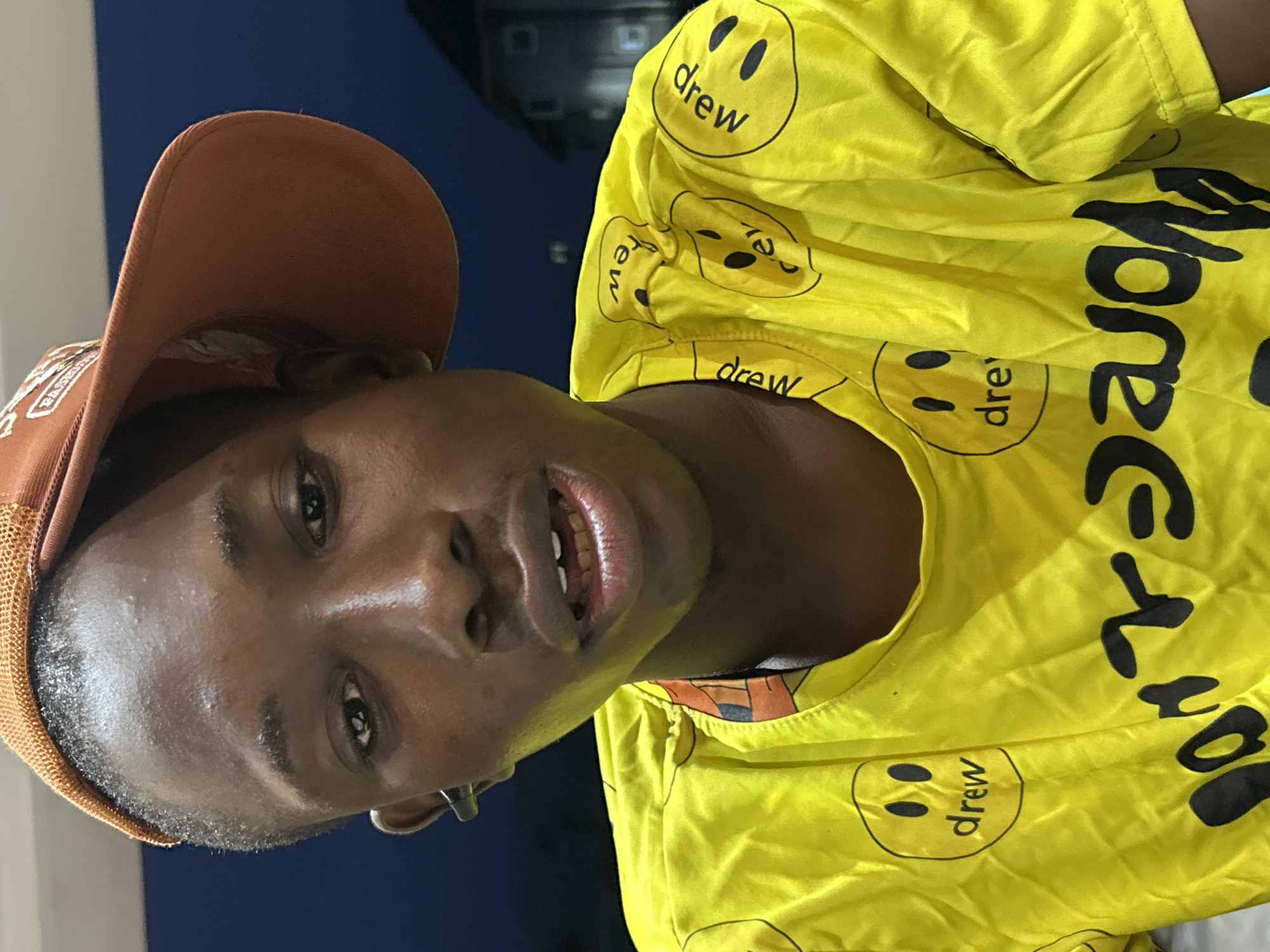 A close-up, self-portrait photograph captures a young Black male wearing a brown baseball cap and a bright yellow t-shirt adorned with black smiley faces and the word "drew". The individual is wearing an earpiece in his left ear. The background is dimly lit and out of focus, suggesting an outdoor setting at dusk or night. The visible portion of the environment includes dark blue tones with faint indications of distant buildings, hinting at a residential or urban area. The specific location of Ihiagwa, Nigeria, is not visually discernible from the image itself.