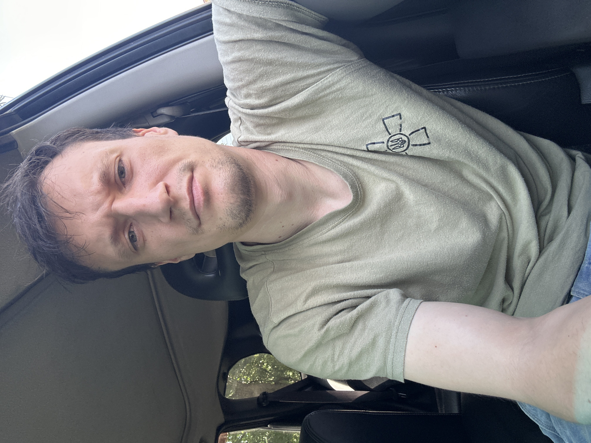 A man with short brown hair and stubble is sitting in the driver's seat of a car. He is wearing a light brown t-shirt with a black emblem on the left side of his chest. The emblem appears to be a stylized cross with a trident in the center. The car's interior is visible, including the roof, a headrest, and the door frame. Through the open car window, trees and foliage can be seen. The scene is consistent with being in Makiv, Ukraine.