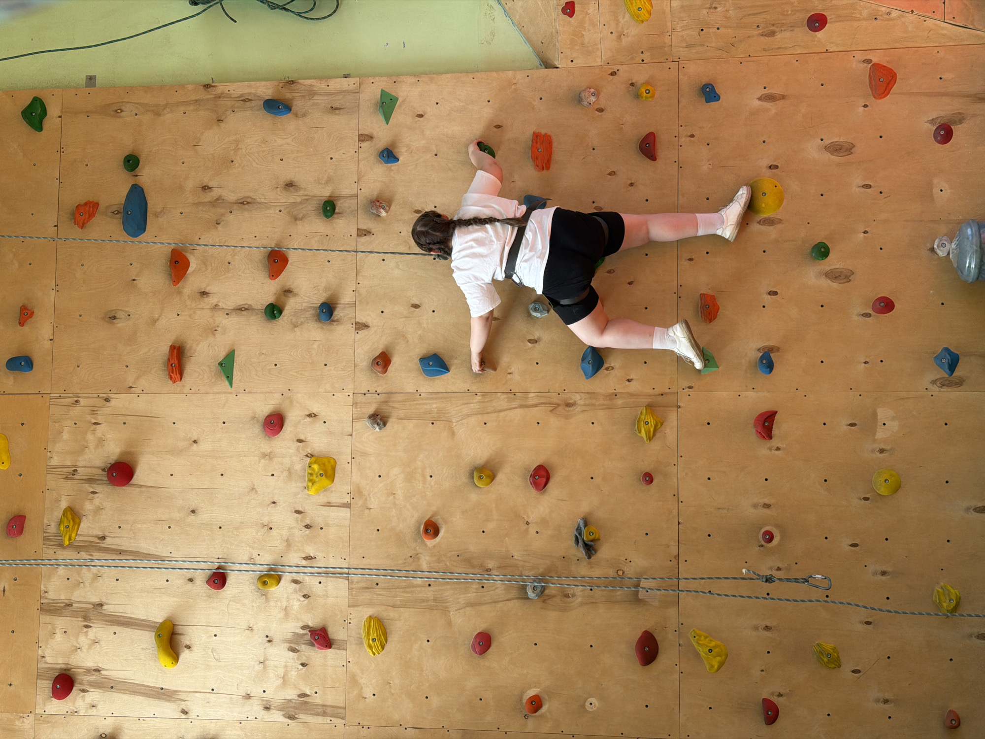 An overhead shot captures a person actively rock climbing indoors on a wooden climbing wall. The climber, who appears to be a young individual with light skin and a single braided ponytail, is dressed in a white short-sleeved t-shirt, black shorts, white socks, and white athletic shoes. They are equipped with a black safety harness around their waist and hips.
The climbing wall is constructed from light-toned wooden panels, arranged in a grid pattern. It is extensively covered with numerous colorful climbing holds of various shapes and sizes, including red, blue, green, orange, yellow, and grey. The climber is positioned horizontally on the wall, stretching their body to reach holds. Their left hand firmly grasps a green hold, while their right arm is extended, seemingly reaching for the next handhold. Their feet are carefully placed on a yellow hold with the left foot and a red hold with the right foot, indicating they are mid-movement during the ascent or traverse.
Two parallel grey ropes span horizontally across the climbing wall. One of these ropes is visibly connected to the climber's harness by a carabiner, suggesting a top-rope climbing setup for safety. In the upper background, a light green wall is visible, with some coiled ropes hanging near the ceiling. A clear plastic water bottle is casually hung on a hold on the far right edge of the climbing wall. The scene depicts an indoor athletic activity within a climbing gym or facility, possibly in Izhevsk, Russia. No visible text is present in the image.