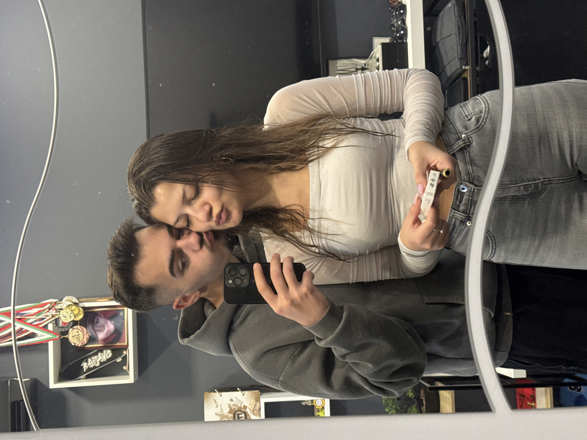 A couple is posing for a selfie in front of a mirror. The woman, on the right, is wearing a white long-sleeved shirt and grey jeans, holding a lipstick and a small white box. The man, on the left, is wearing a grey hoodie and black pants, holding a black smartphone to take the picture. They appear to be kissing or nuzzling close to each other. The background includes a grey wall, a white shelf with various items including medals, a framed picture and a miniature skateboard. The scene appears to be inside a room in Sofia, Bulgaria, based on a sticker of the Bulgarian flag on an item in the background.