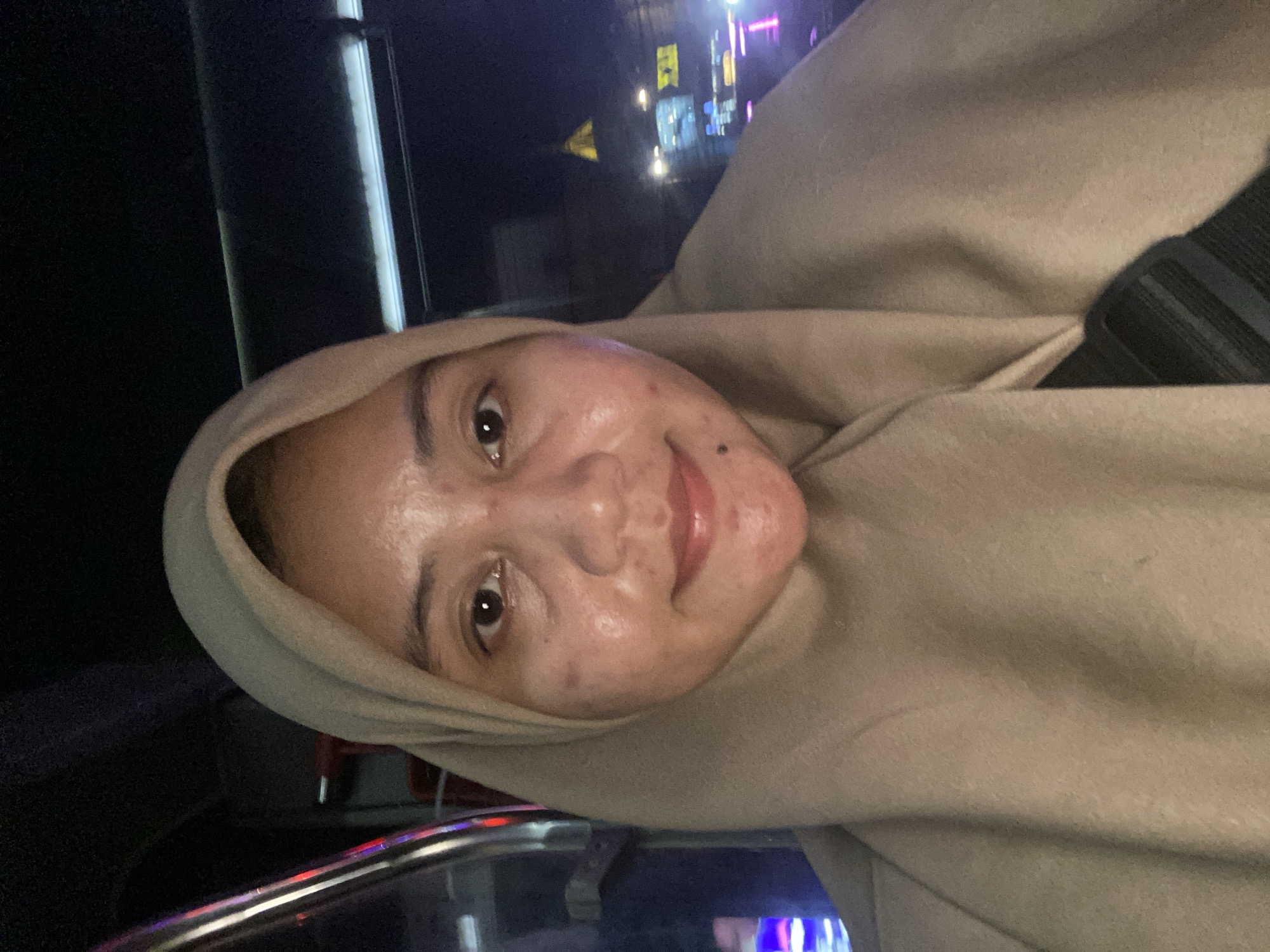 A selfie is taken from a low angle of a young woman wearing a light brown hijab. She is in what appears to be a vehicle at night, with blurred lights and structures visible in the background, suggesting an urban setting. The woman's face is the primary focus, with her eyes looking directly at the camera and a slight smile on her lips. The lighting is dim, with some reflections on her skin, possibly from the ambient light or a flash. The background suggests movement or activity outside the vehicle. The image is not specific enough to determine the exact location within Banjarmasin, Indonesia, but the presence of night lights and vehicle interior implies it was taken in a populated area of the city.