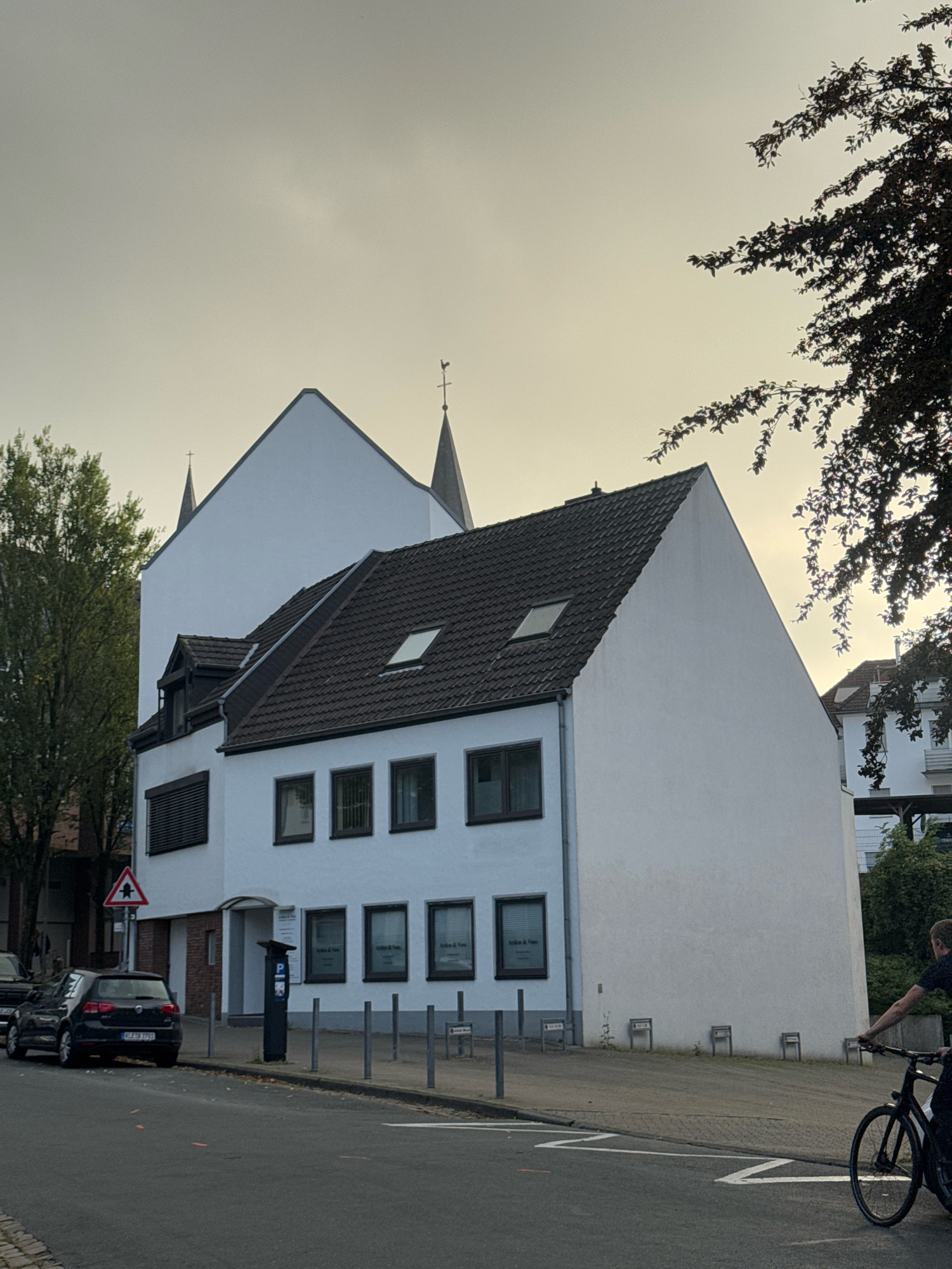 A street-level, eye-level, outdoor, full-shot photograph captures a narrow street in Cleves, Germany. The scene is dominated by a white building with a dark-tiled roof, featuring multiple windows and a steep, gabled section that extends upwards, culminating in a pointed spire topped with a cross and a weather vane. A smaller, dark-roofed extension juts out from the main building, also with windows and a dormer. To the left, a dark-colored hatchback car is parked on the street, and a street sign depicting a pedestrian and a triangular warning symbol is visible. A dark blue parking meter is situated on the sidewalk in front of the building. The street is paved and has white lane markings. A person on a black bicycle is moving from right to left in the foreground. To the right of the building, a portion of another building with a red-tiled roof is visible, along with dense foliage. The sky is overcast and light in color.