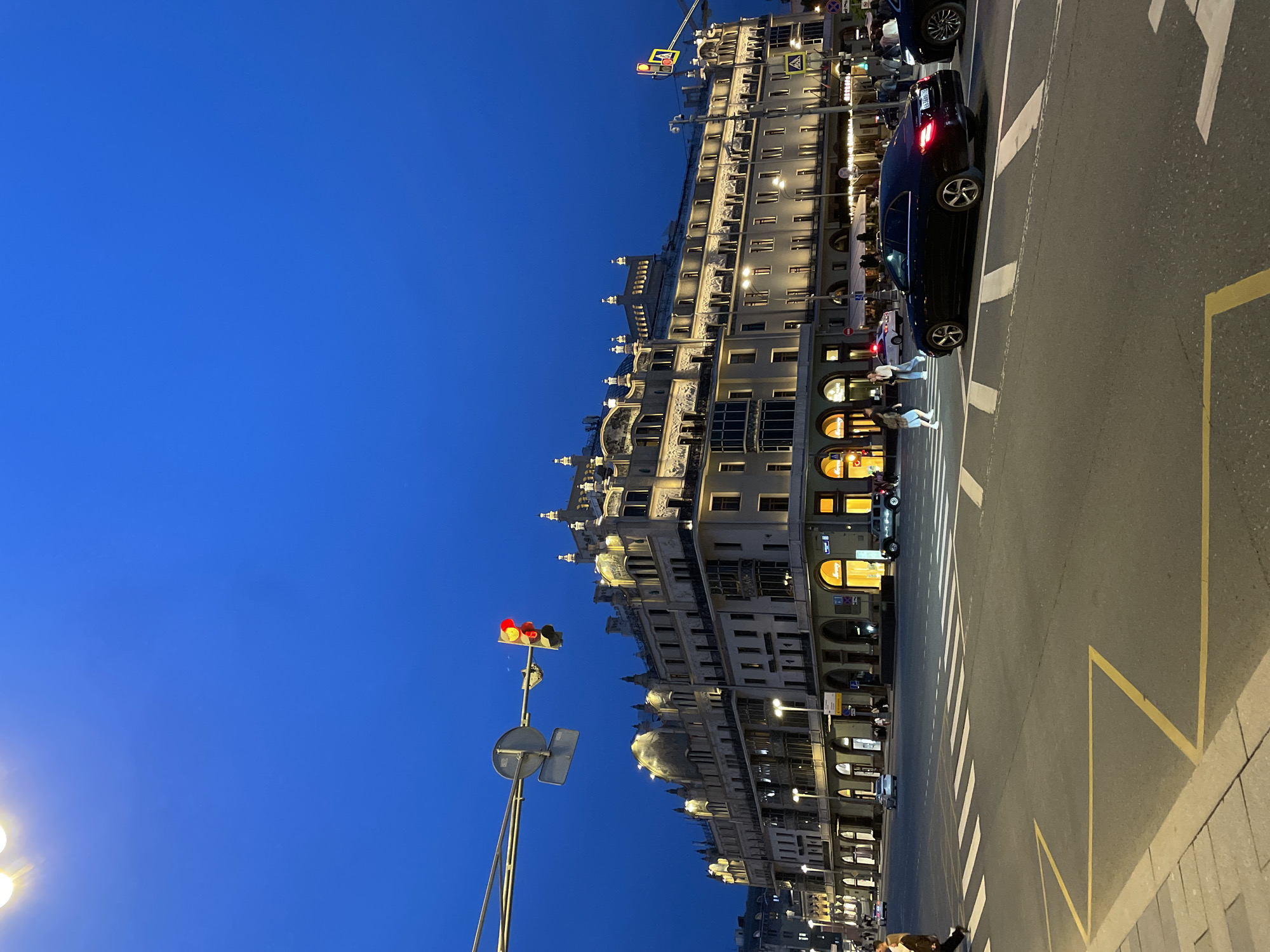 Here's a concise description of the image:
The image depicts a street scene in Moscow, Russia, at dusk or early evening. A large, ornate building with multiple stories and illuminated windows dominates the background. The architecture is classical with arched windows, domes, and decorative elements. A traffic light is visible with a red light illuminated. Pedestrians are crossing the street at a crosswalk. A black car is stopped on the street. The sky is a deep blue color. Road markings and street lights are visible.