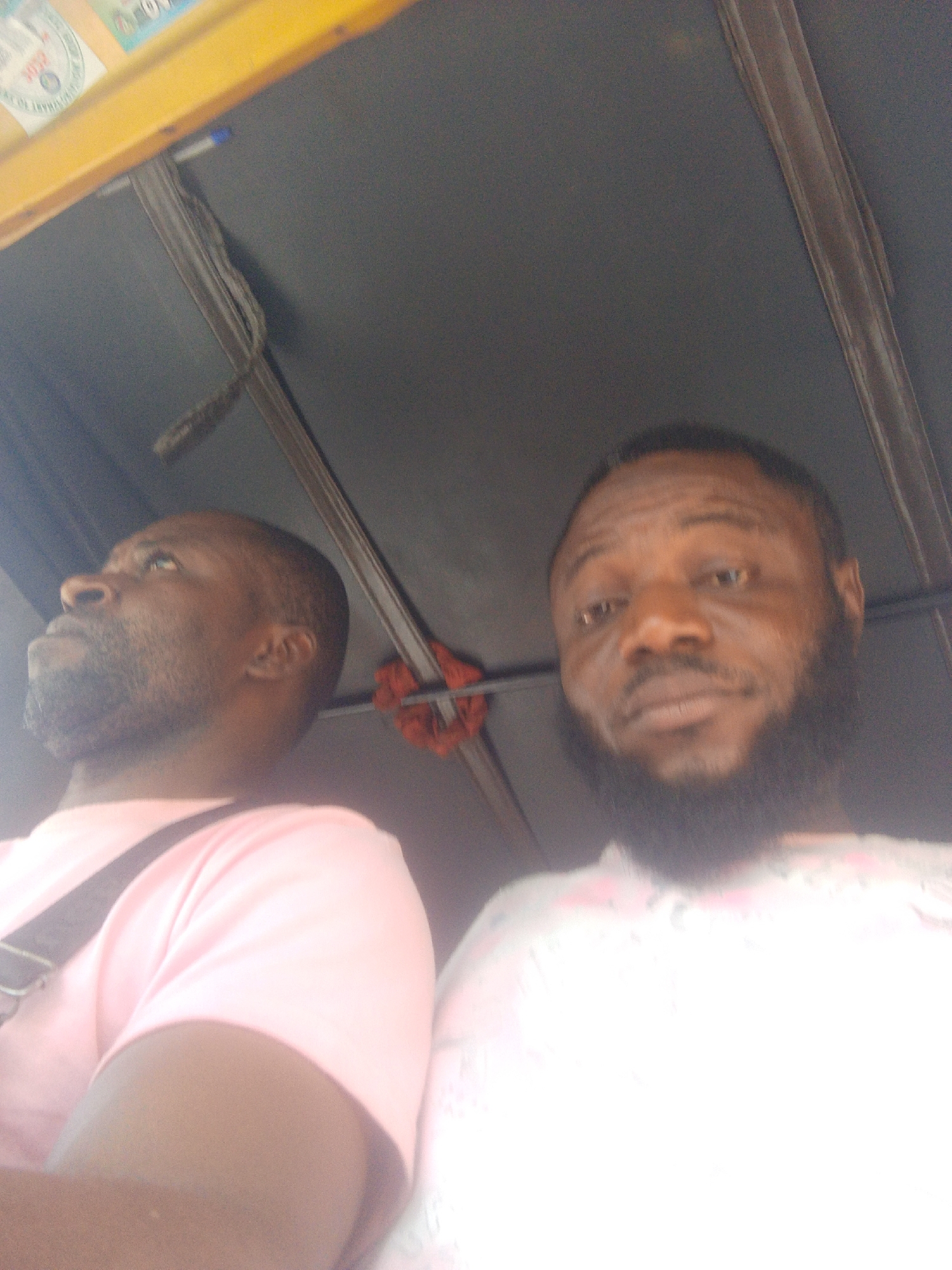 Two dark-skinned men are inside a vehicle, possibly an auto rickshaw, in an urban location. The man on the left, wearing a pink t-shirt, looks upward and has a shoulder strap across his chest. The man on the right has a beard and is looking towards the camera, wearing a light-colored shirt. Above them is the vehicle's roof and frame. The location is Unknown, Unknown.