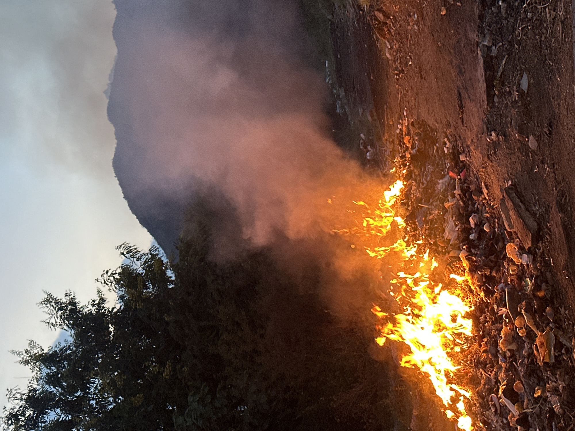 The image shows a fire burning on a dirt slope in Bac Nha Trang Ward, Vietnam. Dense smoke rises from the flames, partially obscuring a tree and a mountain in the background. The sky is visible at the top left, appearing overcast. Debris is scattered on the ground around the fire.