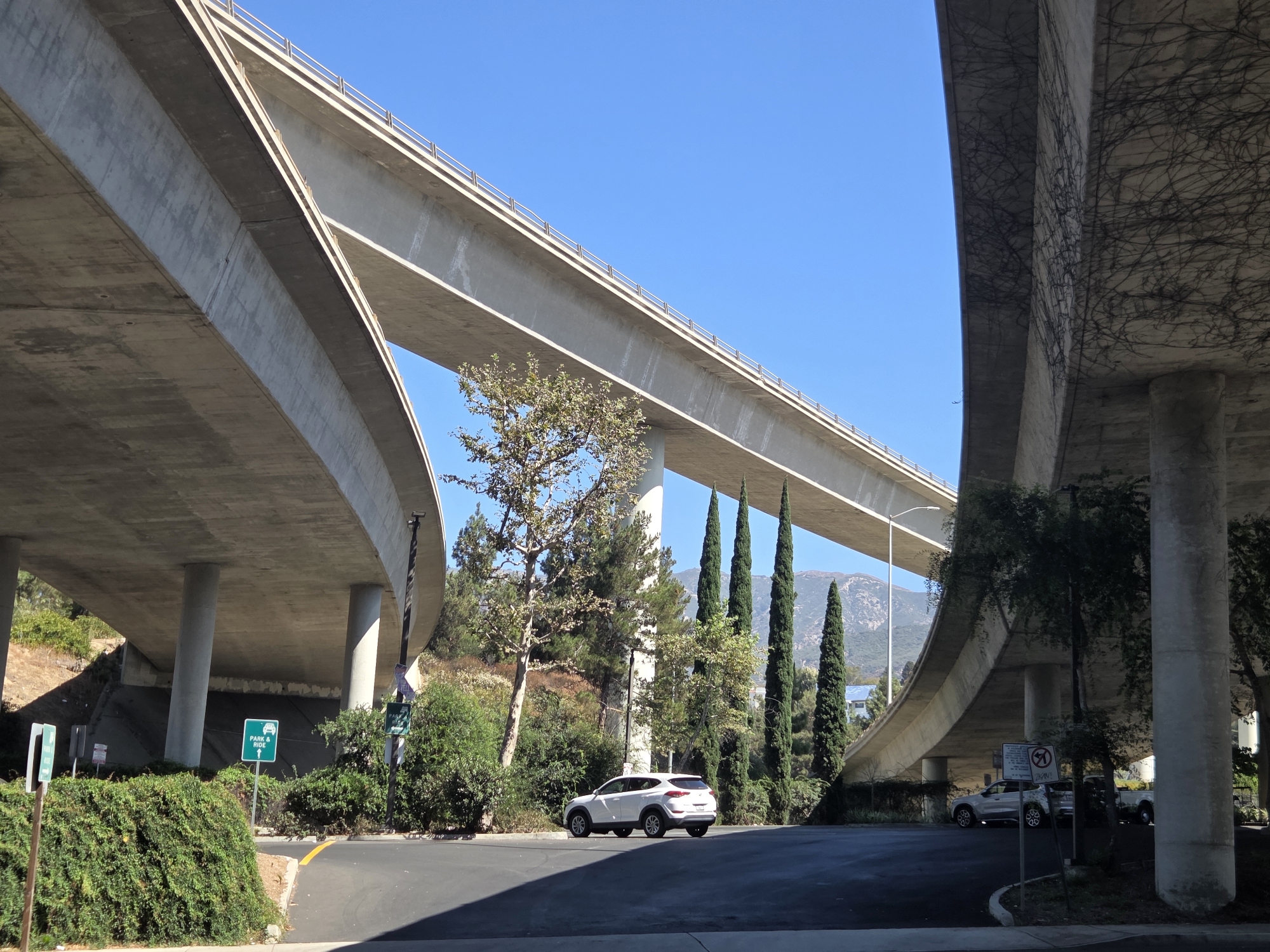 This is a wide-angle, eye-level shot of a concrete overpass in Glendale, California, on a clear, sunny day. The overpass, a significant piece of urban infrastructure, dominates the frame with its imposing concrete structure casting shadows on the ground below. Several massive concrete pillars support the overpass, some of which are partially obscured by lush green foliage and trees. In the mid-ground, a white SUV is parked on an asphalt surface, while in the background, a mountain range is visible under a bright blue sky. The scene is devoid of any people.