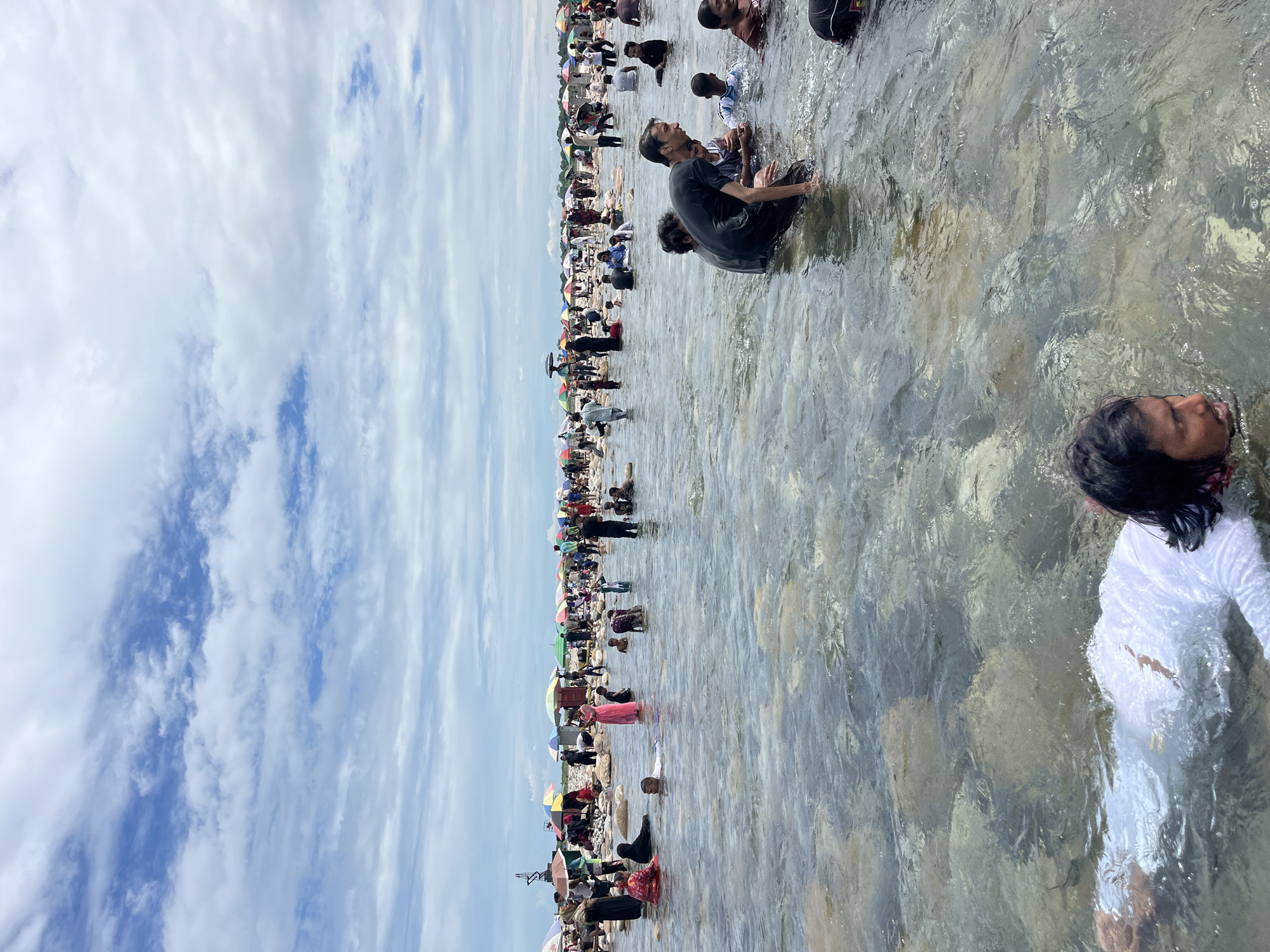 The image depicts a crowded scene at the shores of the River Titas in Majai, India, on a bright, overcast day. A large number of people are wading and swimming in the shallow, rocky river. In the foreground, a person in a white garment is partially submerged and floating on their back. To the right, several men are in the water, some interacting with each other. In the background, a vast crowd of people are gathered on the riverbank, with numerous colorful umbrellas providing shade. The riverbed is visible through the clear water, with large stones and pebbles. The sky is filled with a mix of white and blue clouds.