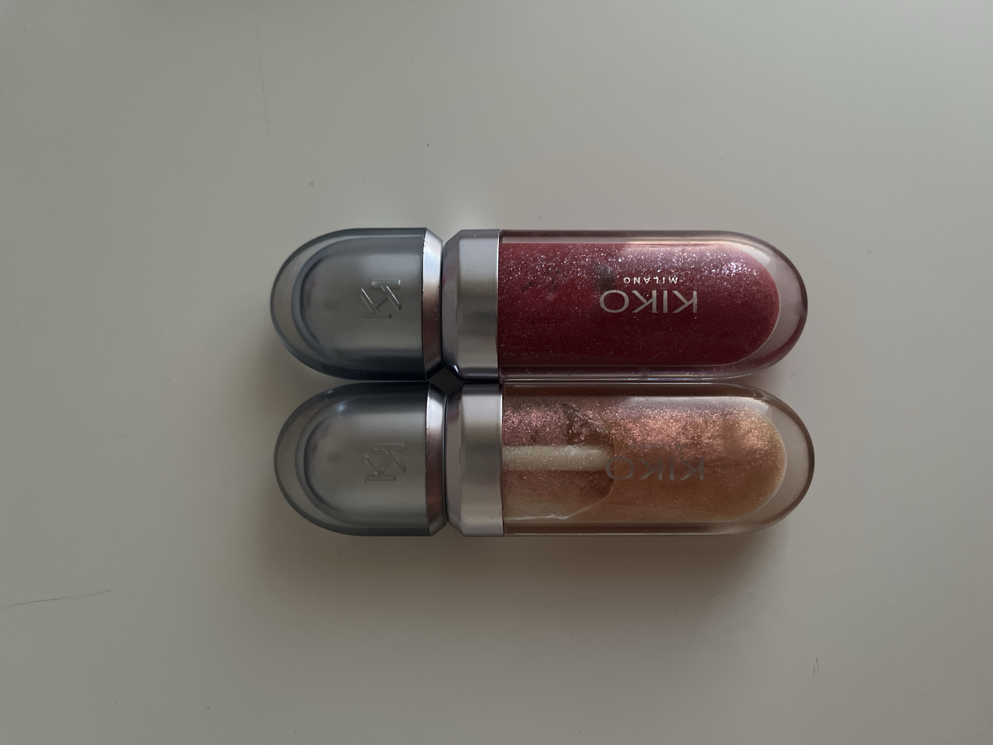 Two Kiko Milano lip gloss bottles are placed side-by-side on a white, flat surface, likely a table or countertop. The bottle on the left contains a shimmering, golden-peach colored gloss, while the bottle on the right holds a vibrant, shimmering red gloss. Both bottles have clear, oval-shaped glass bodies, tapering towards the bottom, and metallic silver caps with the Kiko logo embossed on them. The text "KIKO MILANO" is visible in silver lettering on the glass of the right bottle. The background is a plain, light-colored surface with no discernible details of a city, landscape, or any urban environment. There is no information in the image to suggest the location is Haabneeme alevik, Estonia.