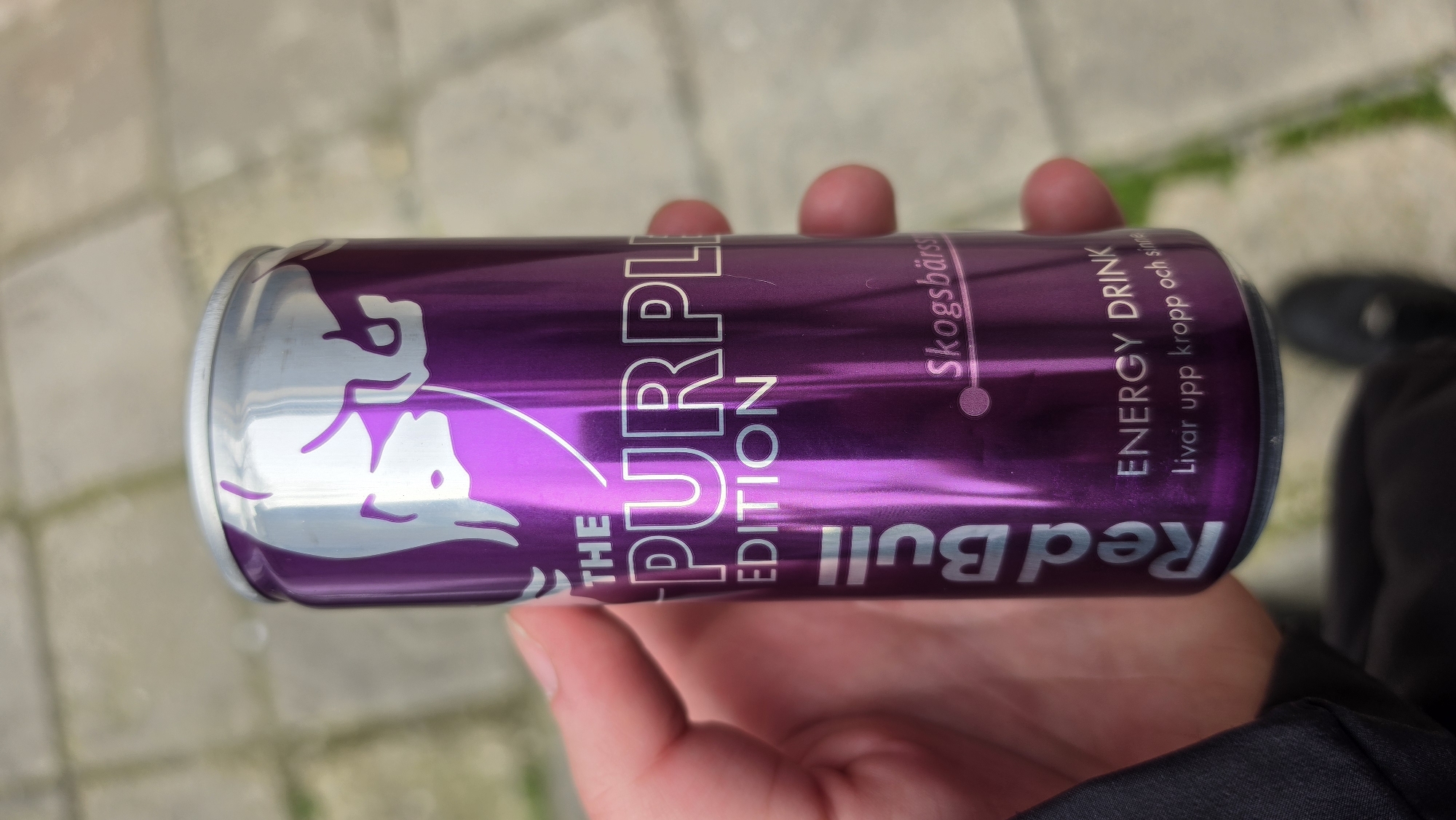 A person's hand holds a purple Red Bull "The Purple Edition" energy drink can. The can is metallic purple with silver logos and text. The text includes "Skogsbärssmak" and "ENERGY DRINK. Livar upp kropp och sinne." The background shows pavement and the edge of a black shoe and dark clothing. The location is not provided in the image.