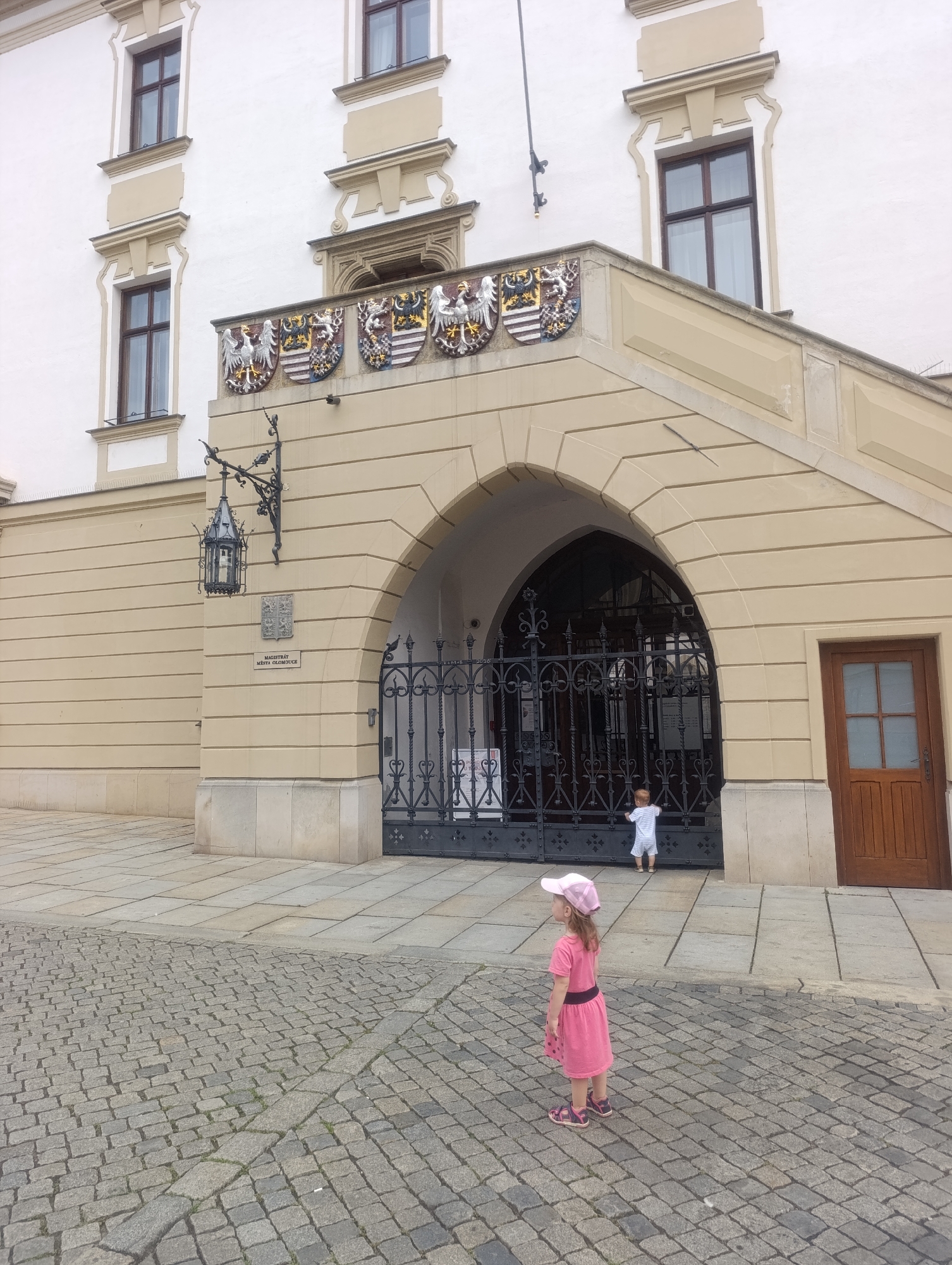 The image depicts two young children standing outside a building with ornate architecture in Olomouc, Czechia. In the foreground, a young girl wearing a pink dress, a pink hat, and sandals stands on a cobblestone path, looking to the left. Behind her, further into the scene, a toddler in a striped outfit stands near a large, decorative metal gate, looking towards it. The building itself is light-colored with large windows, decorative crests above an arched entryway, and a hanging lantern. The scene appears to be set during the daytime, under what seems to be overcast or bright daylight. A sign is visible on the wall near the lantern, and text can also be seen on posters behind the gate. The overall impression is of a family outing at a historic site.