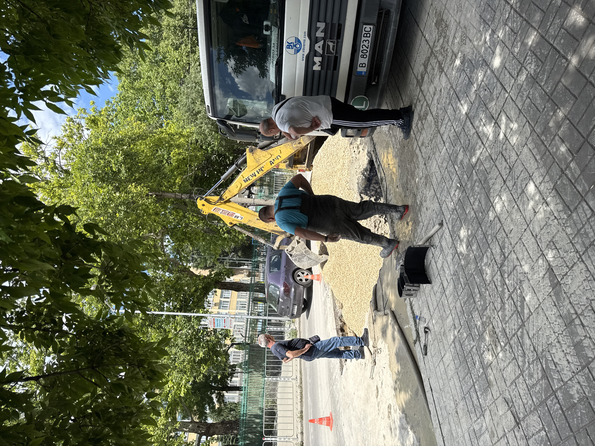 The image depicts a street scene in the city of Varna, Bulgaria, where construction or repair work is underway. In the foreground, a paved sidewalk with a brick-like pattern is visible. Several construction workers are present. One worker, wearing dark overalls and a blue t-shirt, stands with his back to the camera, facing a trench. Another worker, in a white t-shirt and black pants with white stripes, is also facing away, looking towards a truck. A third worker, in blue jeans and a black t-shirt, stands in the street, facing the trench.
A yellow excavator is positioned next to a pile of gravel, with its bucket partially filled with the material. The excavator has markings including "LB959" and "NEW HC APLD". A white truck with the "MAN" logo and license plate "B 8023 BC" is parked nearby, with the website "www.vikvarna.com" displayed on its side. The truck has a logo with "BK" and text in Cyrillic.
The street itself has a trench dug into it, filled with gravel and what appears to be water. A purple car is parked on the street behind the trench. Orange traffic cones are placed on the street to mark the work area. The background shows trees, buildings, and a fence, suggesting an urban environment. The sky is visible, with some clouds. The overall scene suggests an active construction site, likely related to underground utility work.