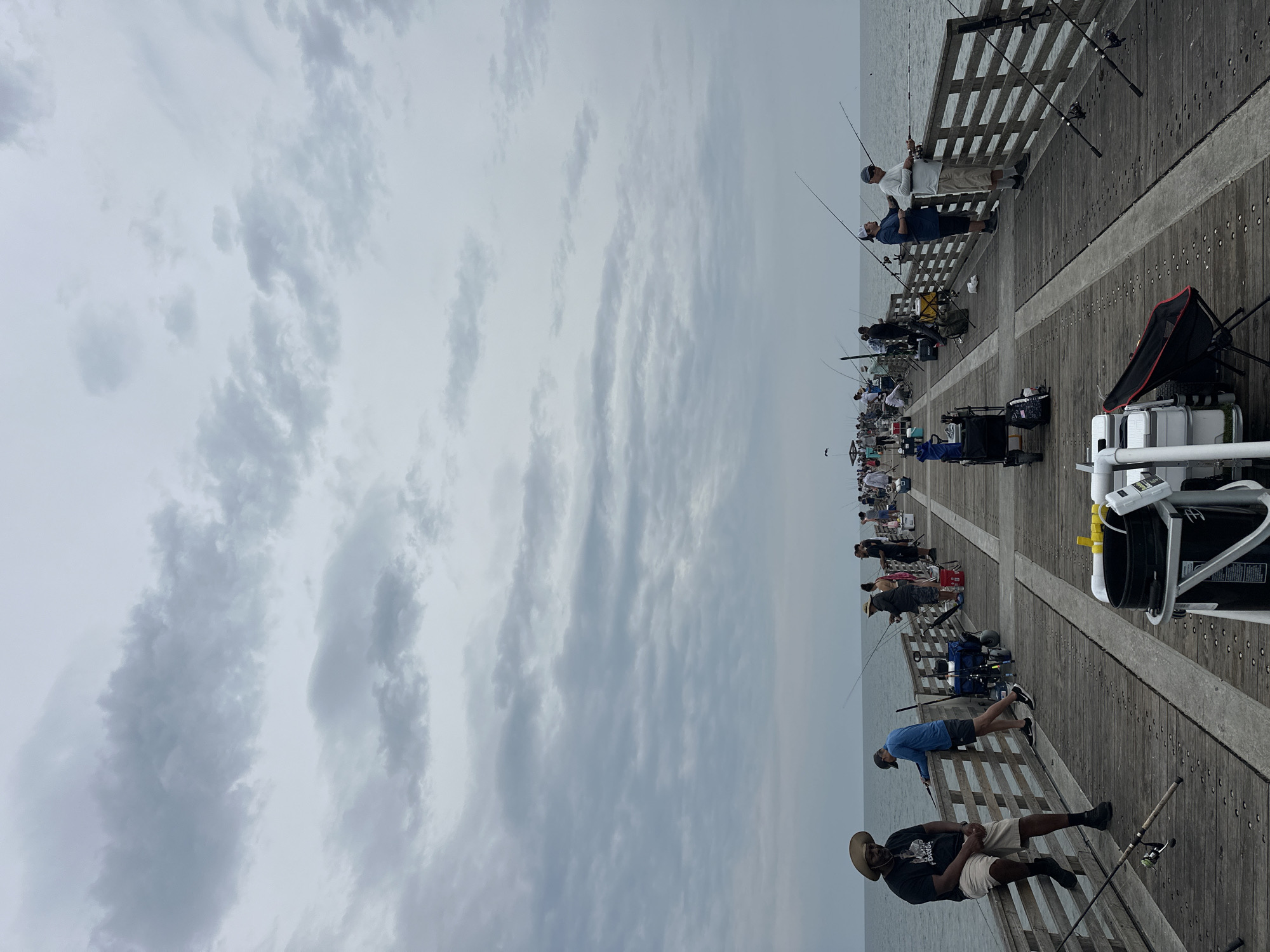 The image depicts a crowded fishing pier at Jacksonville Beach, Florida. Numerous individuals are engaged in fishing, lining both sides of the wooden pier. They are equipped with fishing rods, tackle boxes, and chairs. The sky is overcast with heavy clouds. The pier structure includes wooden planks and railings. The ocean is visible beyond the pier. The scene suggests a popular fishing spot on a cloudy day.