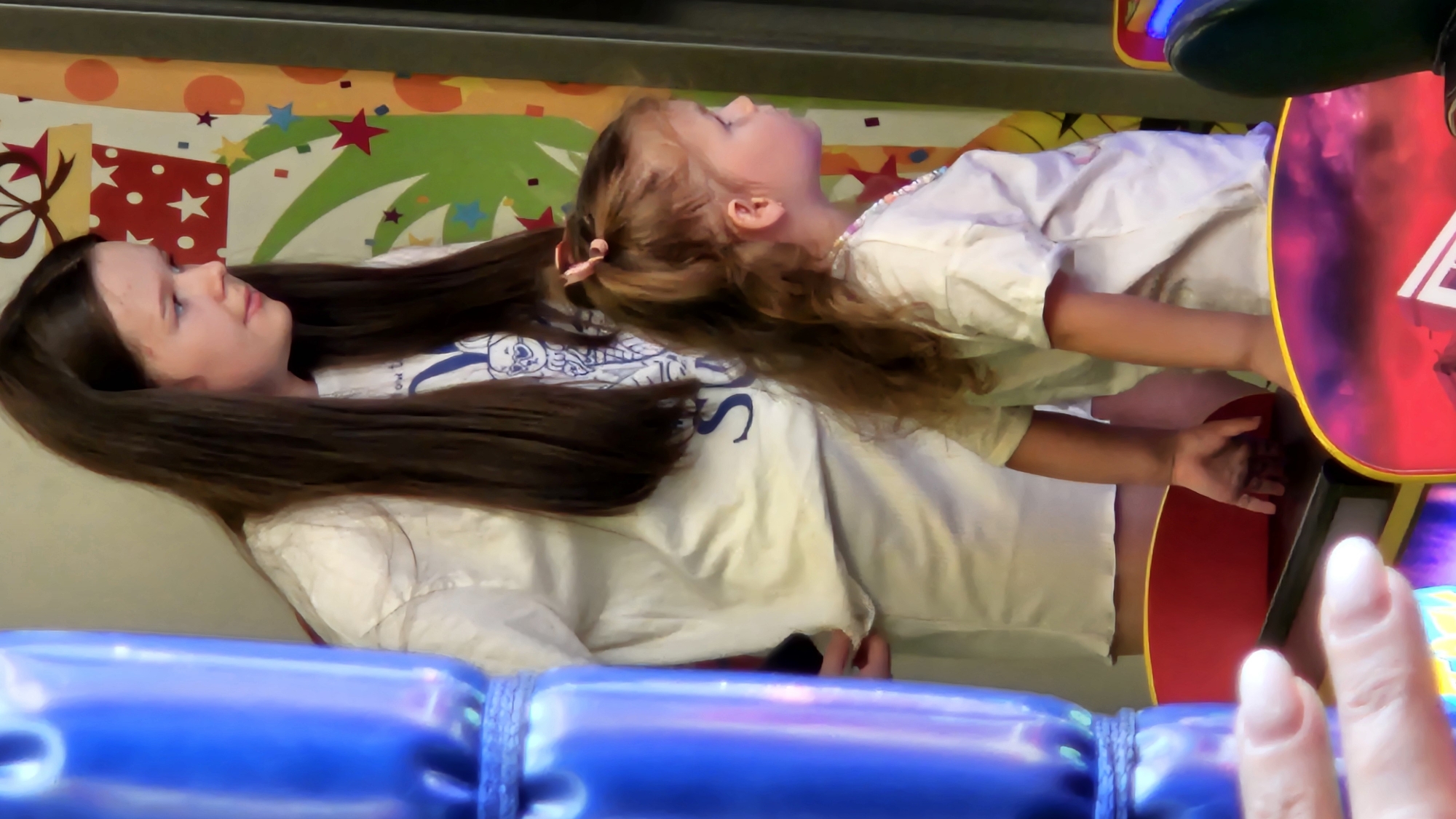 A full-frame, slightly overexposed medium shot captures two young girls in a bright, carnival-like environment. On the left, a tall, slender girl with long, dark brown hair is seen from the chest up. She wears a white t-shirt with a graphic of a bunny on it and has her head turned to the right, looking down and to the side with a slight smile. Beside her, to the right and slightly in front, a younger girl with light brown hair tied in a ponytail with a pink bow is also viewed from the chest up. She wears a white t-shirt and a beaded necklace. Her head is turned to the right, and she is looking intently at something out of frame.
In the foreground, out of focus, a hand with manicured fingernails is reaching towards a black object. The background features a colorful, festive mural with abstract shapes and patterns, suggesting a location like an amusement park or arcade. The overall impression is one of youthful enjoyment and anticipation in a vibrant setting. The image does not contain any discernible elements that would specifically place it in Bratislava, Slovakia.
