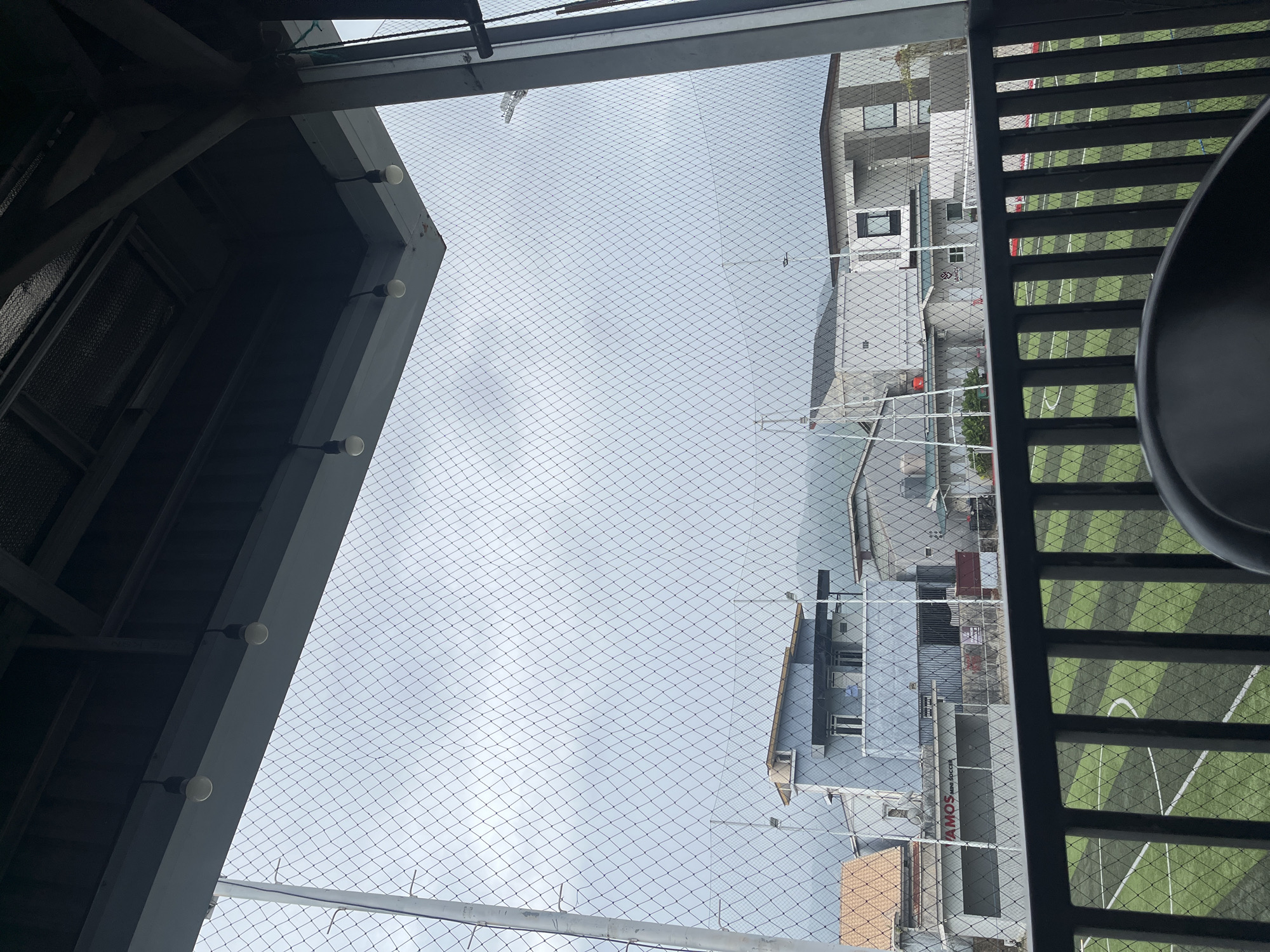 This is a ground-level, eye-level shot taken from a covered balcony or seating area, looking out through a safety net. The foreground features a dark railing and a portion of a dark chair. Beyond the railing, a green and black astroturf soccer field is visible, marked with white lines. A fence separates the field from a row of buildings.

The buildings vary in style and color. One prominent building is a light blue, multi-story structure with several windows and a balcony. Adjacent to it are other buildings, some appearing modern with gray facades and large windows, and one with a reddish-brown tiled roof. In the background, hills or mountains are visible beneath a cloudy, overcast sky. The overall lighting suggests a dim or overcast day. The image is focused on the outdoor scene, with the safety net creating a patterned overlay across the view. The location is identified as Lampeuneurutgampong, Indonesia, and the soccer field appears to be part of a sports facility. No people are clearly visible in the scene.