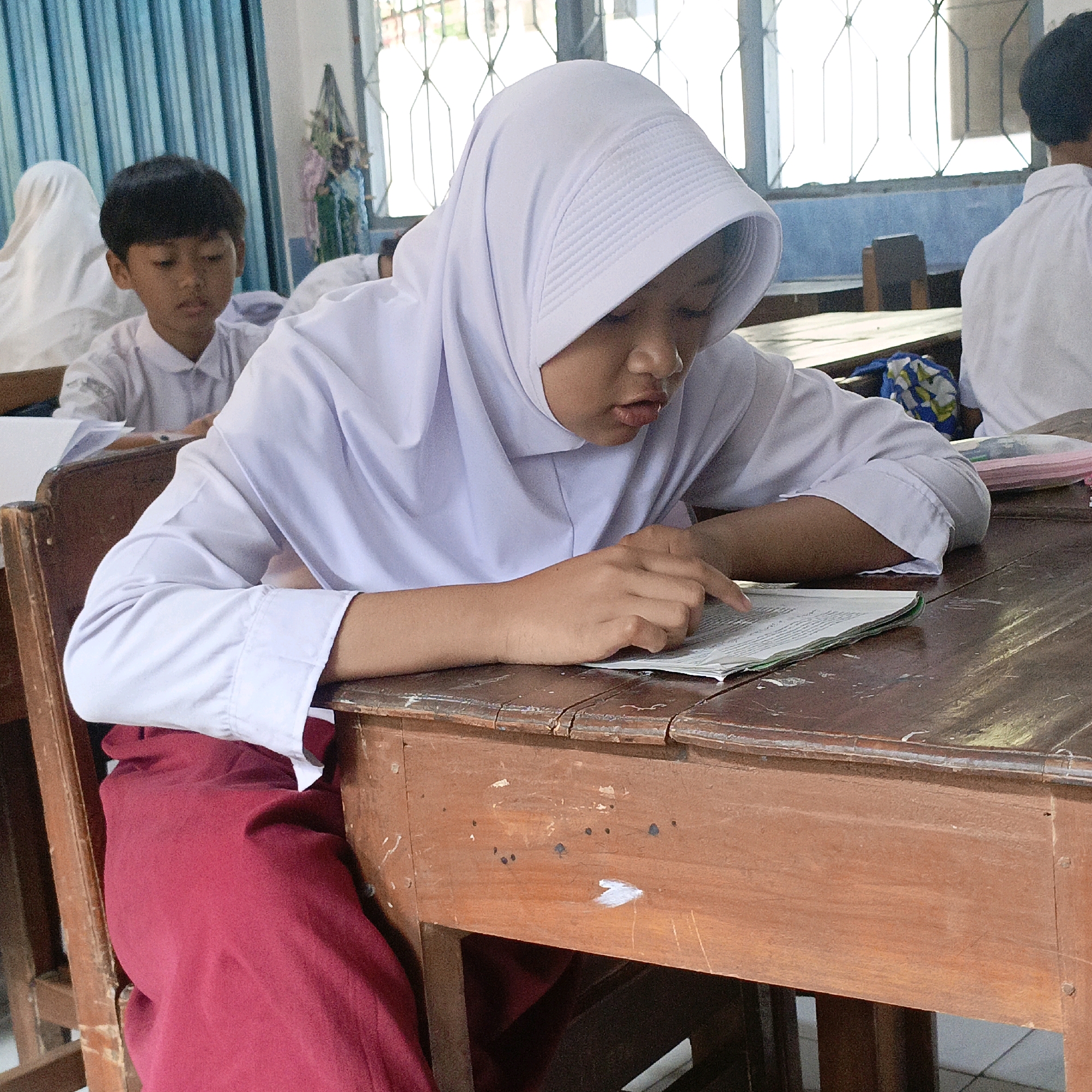 In a classroom setting in Salatiga, Indonesia, a young girl wearing a white hijab and a white school uniform with a red skirt is seated at a wooden desk, engaged in reading a book. Her right hand is resting on the open book, with her index finger pointing to a line of text, suggesting she is actively studying or following along. To her left, another student, a boy, is also seated at a desk and appears to be looking down. In the background, other students and parts of the classroom, including desks and windows with blue curtains, are visible. The overall atmosphere suggests a typical school day, with students focused on their learning activities.