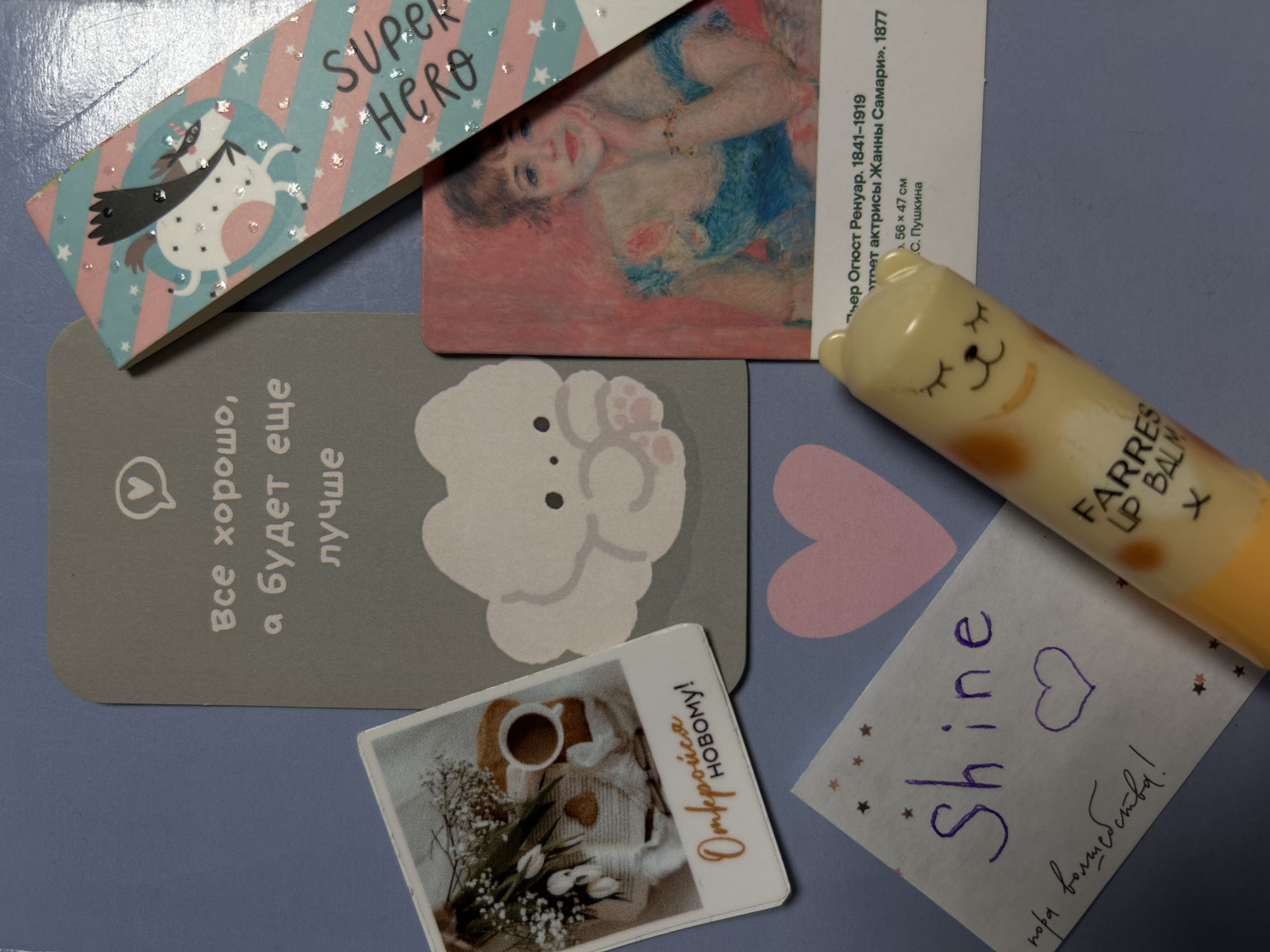 The image presents a flat lay arrangement of various cards, a lip balm, and a handwritten note on a blue-grey surface.
In the top left, a light blue and pink striped bookmark or small card features the English text "SUPER HERO" with glitter stars. It depicts a whimsical, unicorn-like creature wearing a dark cape and sparkling spots.
To its right is a reproduction of a painting, likely a postcard. Visible Russian text identifies it as "Пьер Огюст Ренуар. 1841-1919. Портрет актрисы Жанны Самари». 1877 г." (Pierre-Auguste Renoir. 1841-1919. Portrait of actress Jeanne Samary. 1877.), along with dimensions "56 х 47 см" and "С. Пушкина", implying it's associated with the Pushkin State Museum of Fine Arts. The painting shows a woman with reddish-brown hair, wearing a blue dress, looking directly at the viewer.
Below the "SUPER HERO" card is a grey card featuring a white cloud-like creature with prominent paws. Russian text is vertically oriented, stating: "Все хорошо, а будет еще лучше" (Everything is good, and it will be even better).
Near the bottom center, a small card displays a photograph of a cup of coffee, white tulips, and baby's breath flowers on a light-colored woven surface. Golden Russian text on the card reads: "Открой новому!" (Open to new!).
A yellow lip balm, designed with a cat face (closed eyes), lies diagonally across the right side of the image. Its label is partially visible, showing "FARRES LIP BALM".
Beneath the lip balm, a small white paper has hand-written text. The English word "Shine" is written in purple, accompanied by a purple heart outline. Below it, in cursive, is the Russian phrase "пора волшебства!" (time for magic!). A pink heart cut-out is visible on the blue-grey surface beneath this note.
The collection of items, including the art reproduction from the Pushkin Museum and the various pieces with Russian text, aligns with the given location context of Saint Petersburg, Russia. The overall arrangement suggests themes of inspiration, art appreciation, and personal well-being.