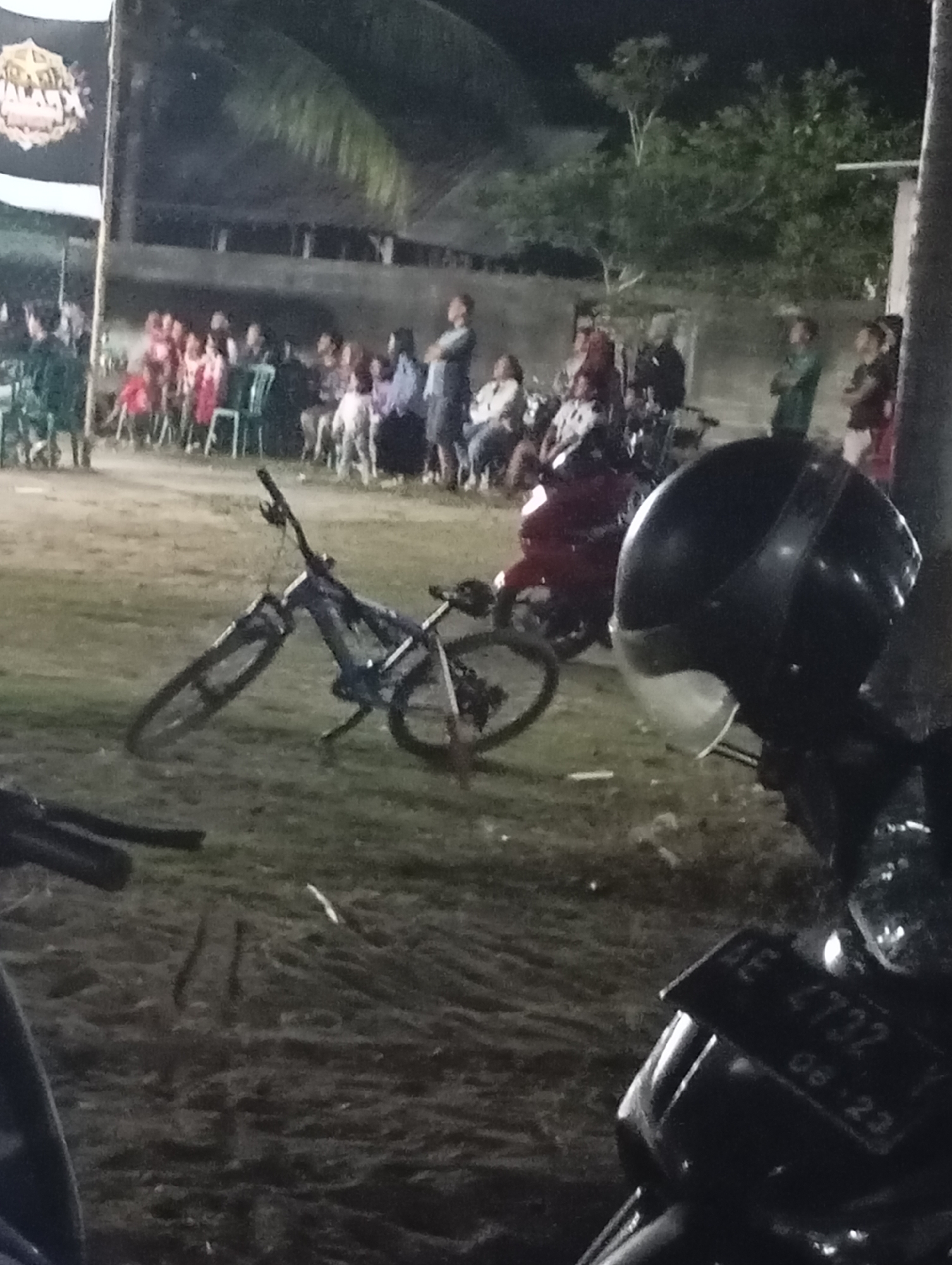 The image depicts a nighttime outdoor gathering in Pacitan, Indonesia. In the foreground, a black helmet sits on a motorcycle. To the left, a bicycle is propped up on its kickstand. In the background, a crowd of people is seated and standing, appearing to watch an event. The environment is dimly lit, suggesting it is evening or night. The overall scene captures a community event or festival in a semi-rural or suburban setting within Pacitan.