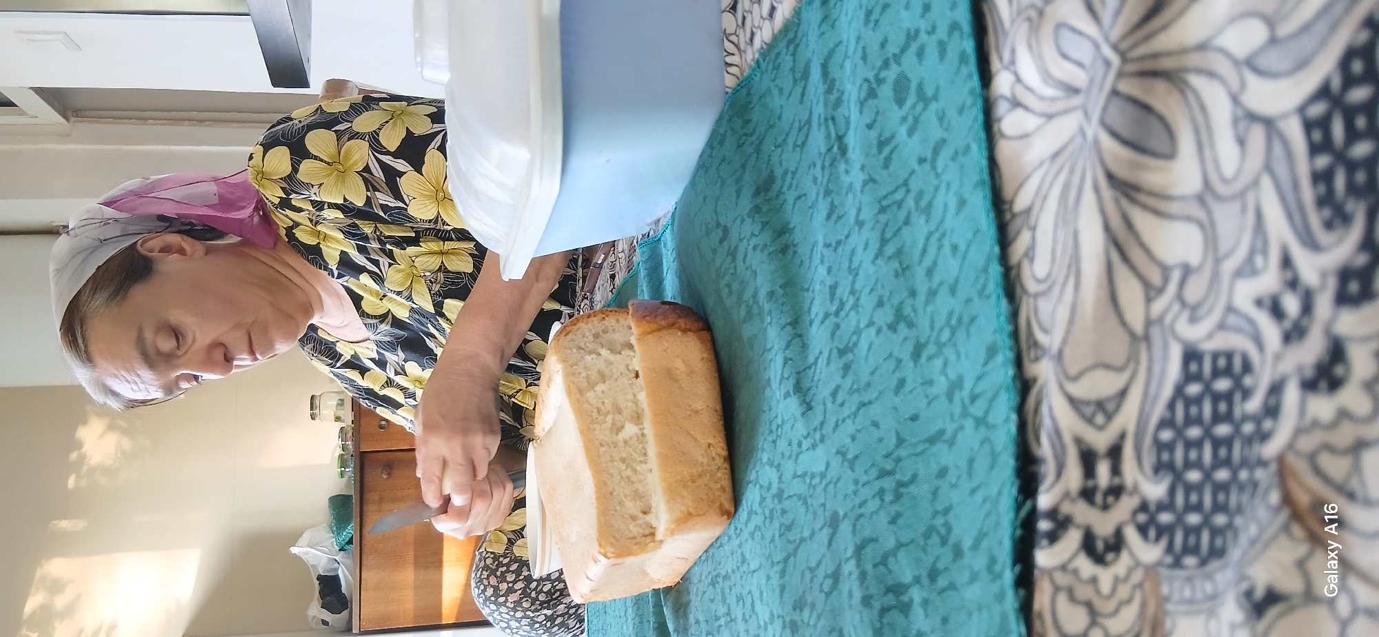 In Angren, Uzbekistan, a woman with graying hair, wearing a headscarf and a yellow and black floral print dress, is sitting at a table and preparing food. She holds a knife in her right hand, with her left hand supporting a small piece of food, possibly cheese or butter, which she is about to cut. In front of her on a teal-colored tablecloth is a loaf of bread, partially sliced, and a light blue plastic container with a white lid. The background shows a wooden cabinet with jars and bags on top, and a wall with shadows cast on it. The scene is indoors, likely in a home kitchen or dining area.