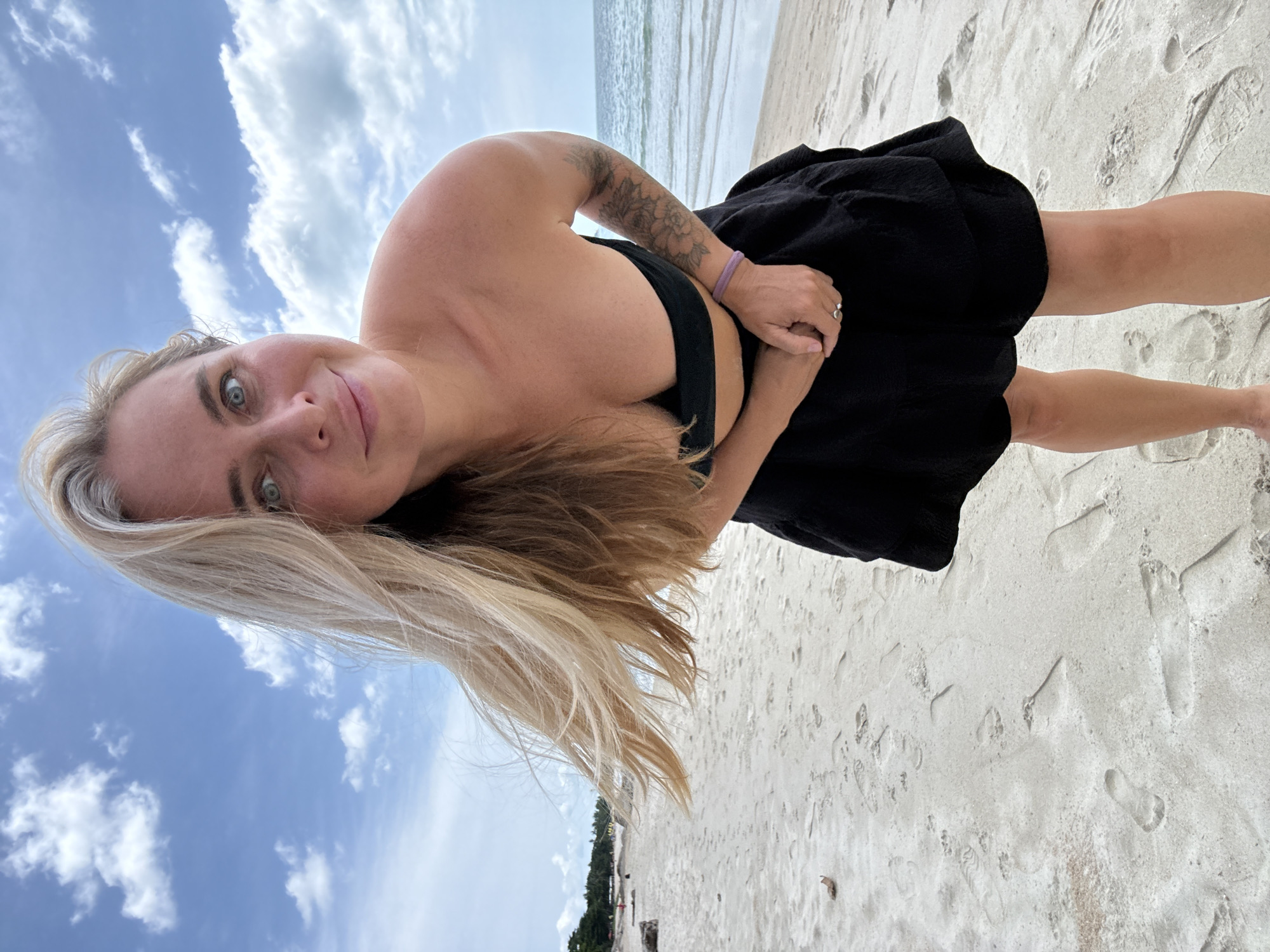 A woman with blonde hair and blue eyes is posing on a sandy beach in Ko Samui District, Thailand. She is wearing a black bikini top and a black skirt. Her hair is blowing in the wind. The ocean is visible in the background. The sky is blue with some clouds. There are footprints in the sand.