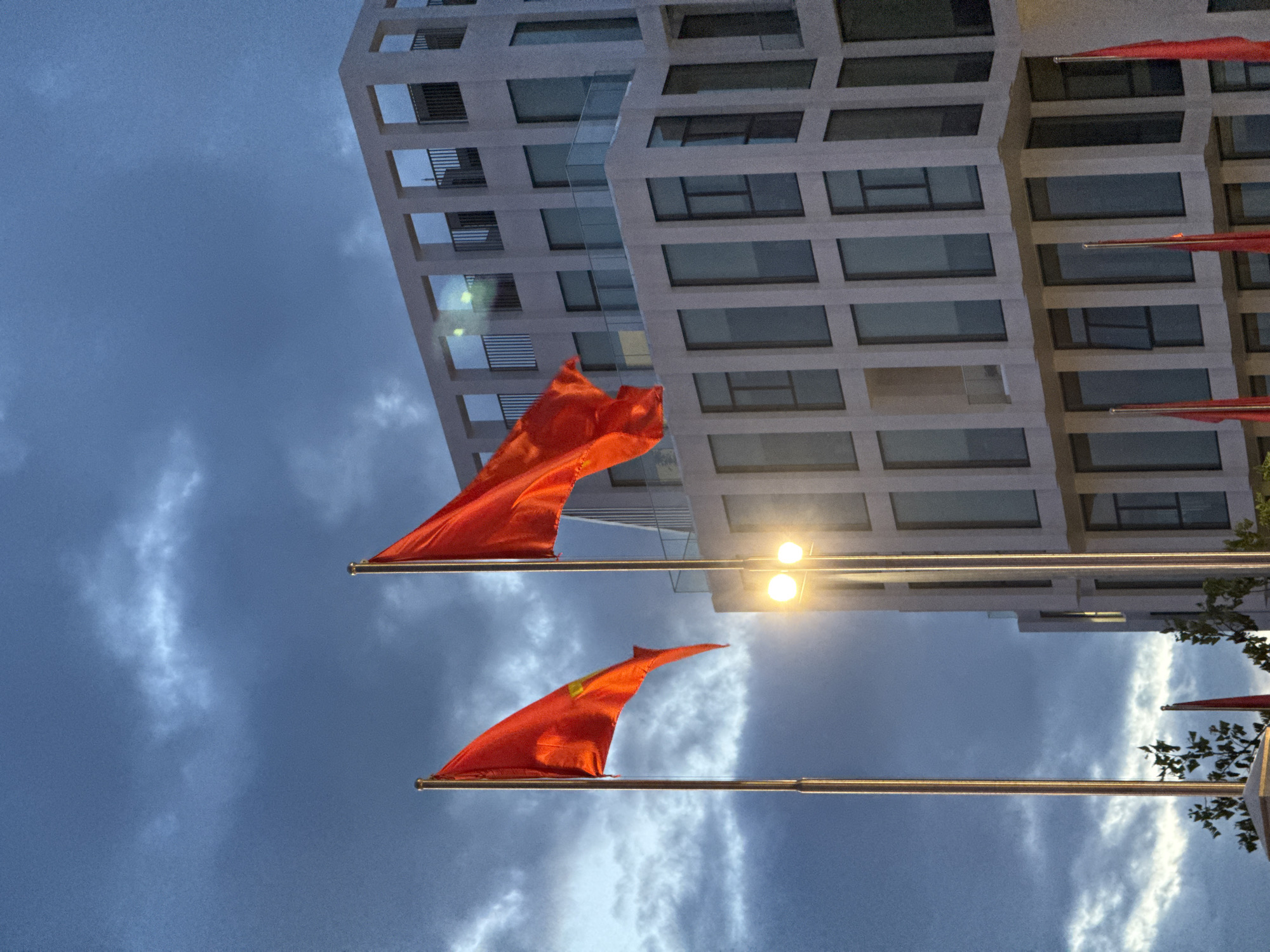 The image depicts two red flags with yellow stars, fluttering in front of a modern white building with a grid-like facade and multiple windows. The sky above is overcast with dramatic clouds, suggesting twilight or stormy weather. A street lamp with two bright lights is positioned between the flags. In the background, there are hints of greenery and other red flags. This scene is located at the Square of Two September, a prominent public space in Nha Trang, Vietnam, known for its flags and adjacent modern architecture.