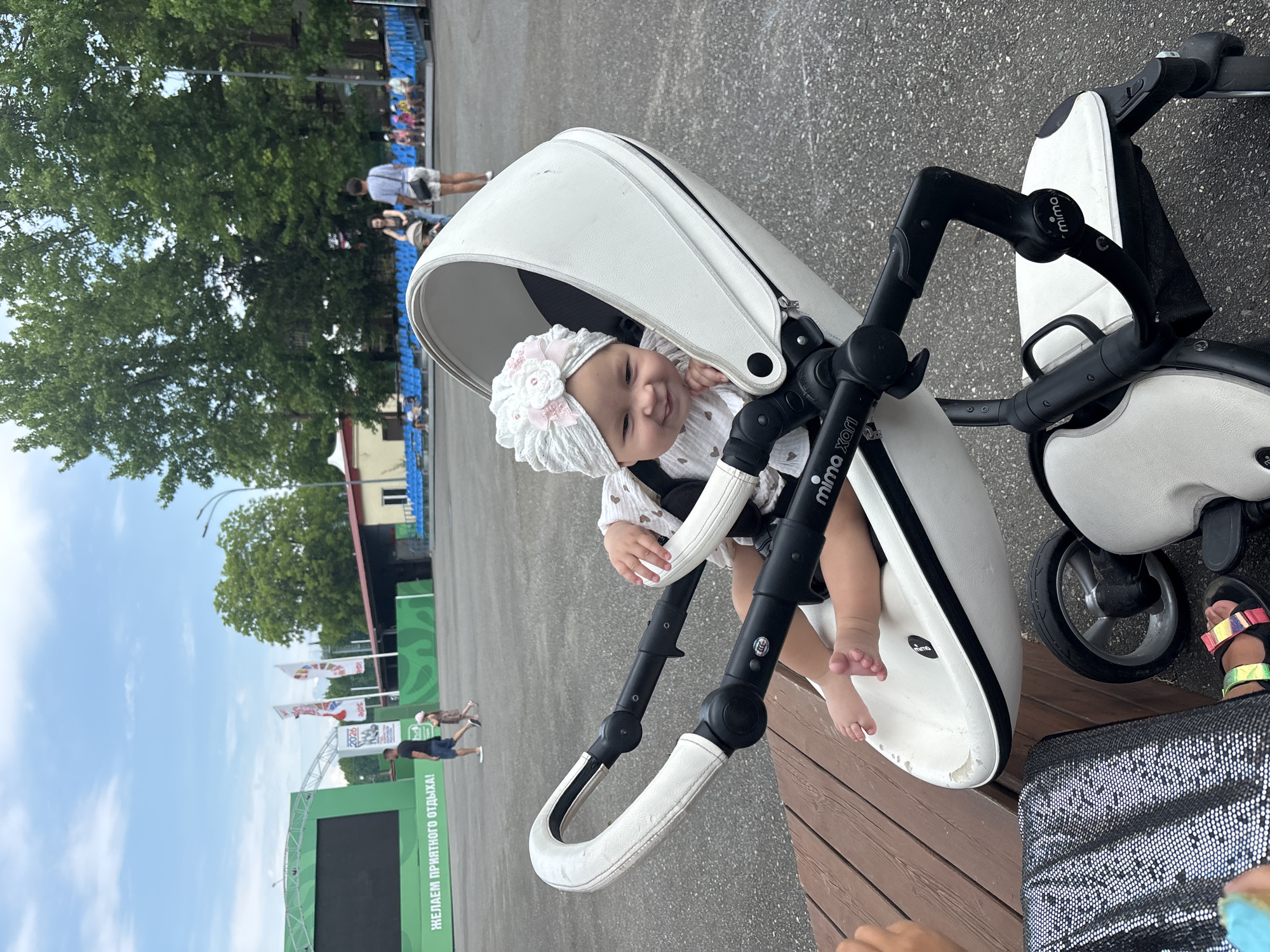 The image shows a baby in a white stroller. The baby is wearing a white hat with pink flowers and a white outfit. The stroller is modern and sleek, with black accents. The background is blurred but appears to be an outdoor setting with trees and a blue sky. There are some people in the background, and what looks like a stage or event setup. One of the crops shows a sign with Russian text that reads "ЖЕЛАЕМ ПРИЯТНОГО ОТДЫХА!", which translates to "We wish you a pleasant rest!". The image appears to be taken during the day, with bright lighting. The overall mood is cheerful and relaxed, with the baby looking happy and content. The location is identified as Sochi, Russia, suggesting this might be during a family outing or event in the city. The stroller has "Mima Xari" branding visible on it.