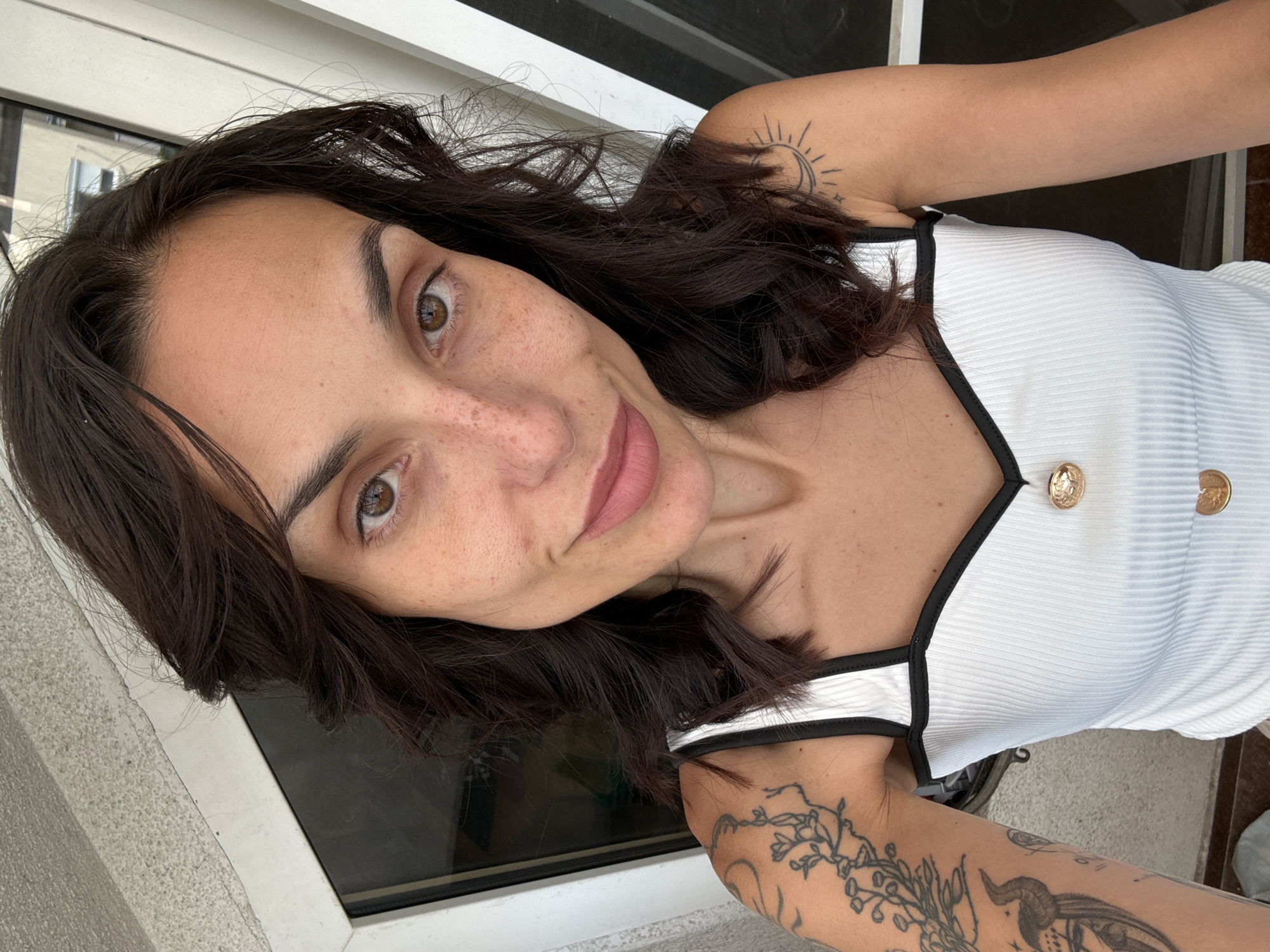A medium shot portrait shows a woman with dark brown hair and brown eyes, looking directly at the camera. She has a light complexion with freckles across her nose and cheeks. Her lips are slightly parted and appear to be painted with a nude lipstick. She is wearing a white ribbed tank top with black trim and gold buttons. Tattoos are visible on her left arm and right shoulder. The background appears to be an outdoor setting, possibly a balcony, with a window or glass door behind her. The lighting suggests it is daytime. No specific landmarks or details indicative of Pleven, Bulgaria are present in the image.