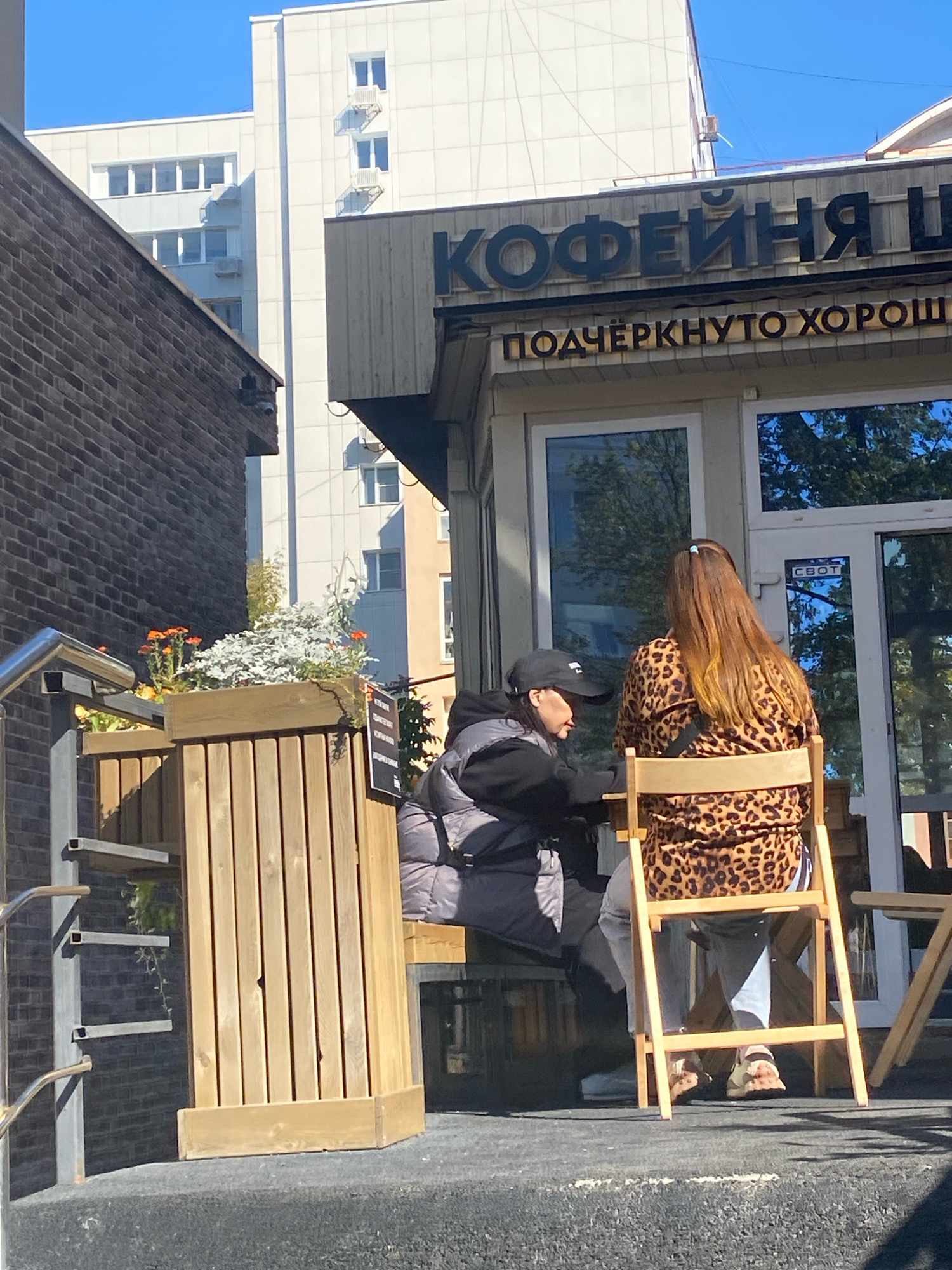 The image depicts an outdoor seating area of a cafe in Perm, Russia. Two women are seated at a table. One woman, wearing a black baseball cap and a black puffer vest over a black hoodie, is looking down towards the table. The other woman, seated in a wooden chair and facing away from the camera, is wearing a leopard-print blouse and light-colored pants. Her feet, clad in sandals, are visible. Behind them, a building with large windows and signage in Russian reading "КОФЕЙНЯ Ц" and "ПОДЧЁРКНУТО ХОРОШО" (Coffee shop C and Emphatically Good) is visible. A brick building is situated to the left of the cafe, and a modern apartment building with a white facade is in the background. A wooden planter box with flowers and foliage is positioned in the foreground to the left. The scene is illuminated by natural daylight, suggesting it is daytime.