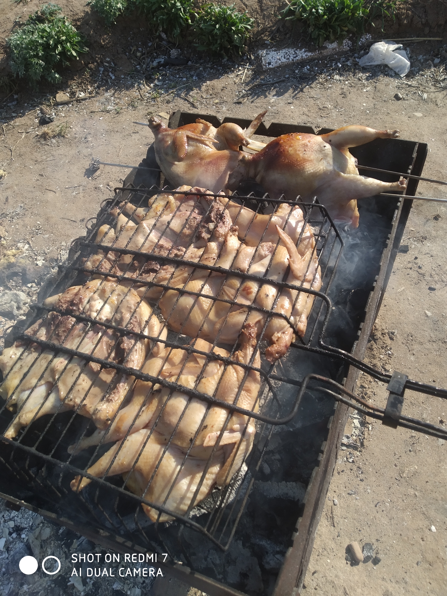 The image depicts a barbecue scene. Two whole chickens are roasting on spits over a charcoal grill. In the foreground, a metal grill holds pieces of chicken, also being cooked. Smoke rises from the grill, indicating active cooking. The barbecue is located outdoors on a dirt or sandy surface, with some vegetation visible in the background. A plastic bag is visible next to the grill. The photograph was taken with a Redmi 7 AI Dual Camera. Although the location is requested to be Chaikovsky, Russia, there is no evidence within the image to confirm this location.