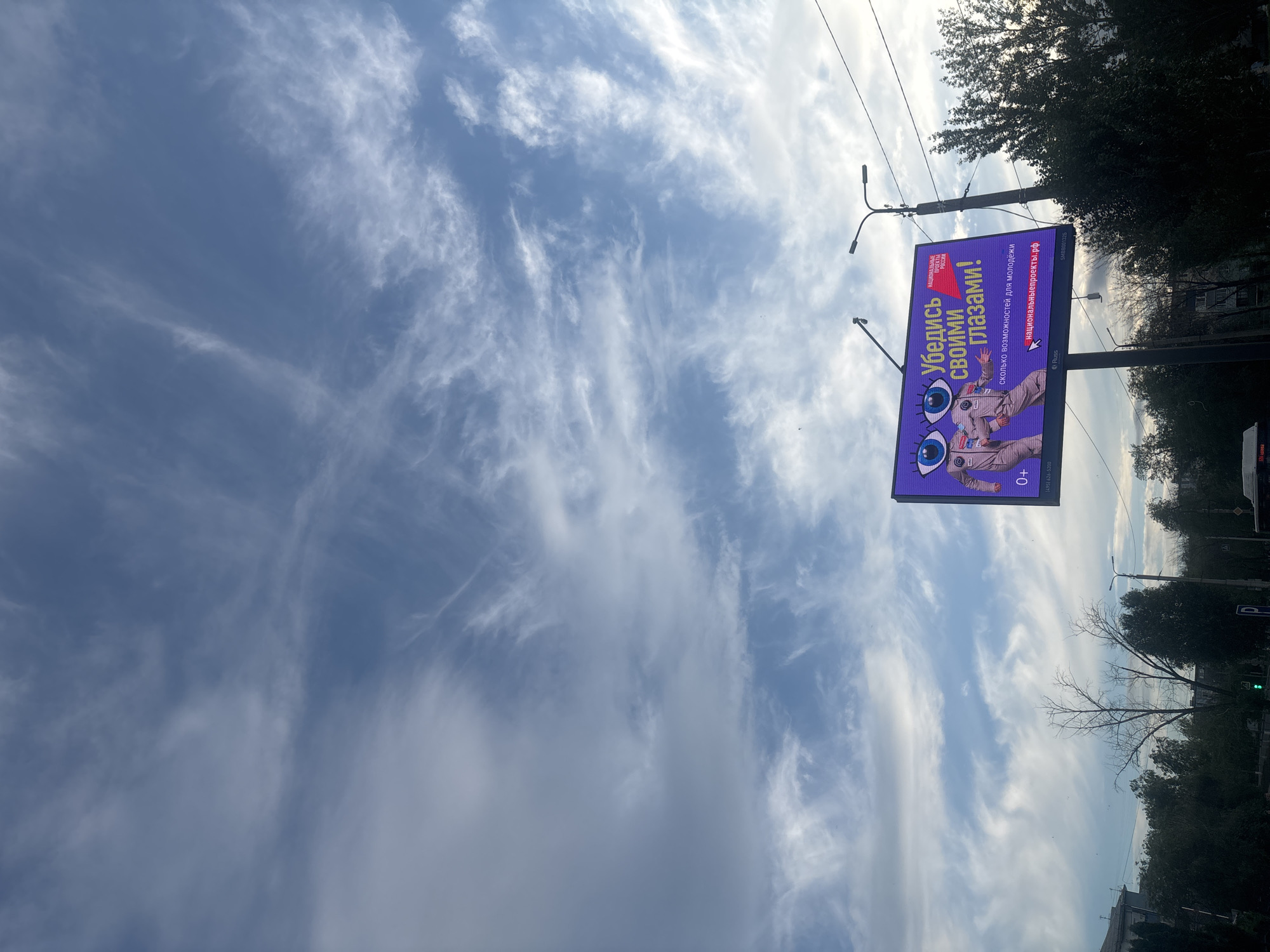 The media file shows a billboard against a backdrop of a partly cloudy sky. The billboard features a cartoonish image of a person in a spacesuit with large, wide-open eyes. Text on the billboard in Russian reads "Убедись своими глазами!" which translates to "See for yourself!". Below this, it says "сколько возможностей для молодёжи" meaning "how many opportunities for youth". The website "национальныепроекты.рф" is also visible, which translates to "nationalprojects.rf". The age rating "0+" is displayed in the bottom right corner of the billboard.
The scene is set outdoors, with trees visible along the right side of the image, suggesting a street or park. Streetlights are also present. The sky is filled with wispy cirrus clouds, indicating a day with variable cloud cover. The lighting suggests it is daytime, possibly late afternoon or early evening due to the angle of the light filtering through the trees and clouds. The overall mood conveyed by the billboard image is one of wonder and opportunity, emphasized by the large, surprised eyes. The context provided states the location is Novokuybyshevsk, Russia.