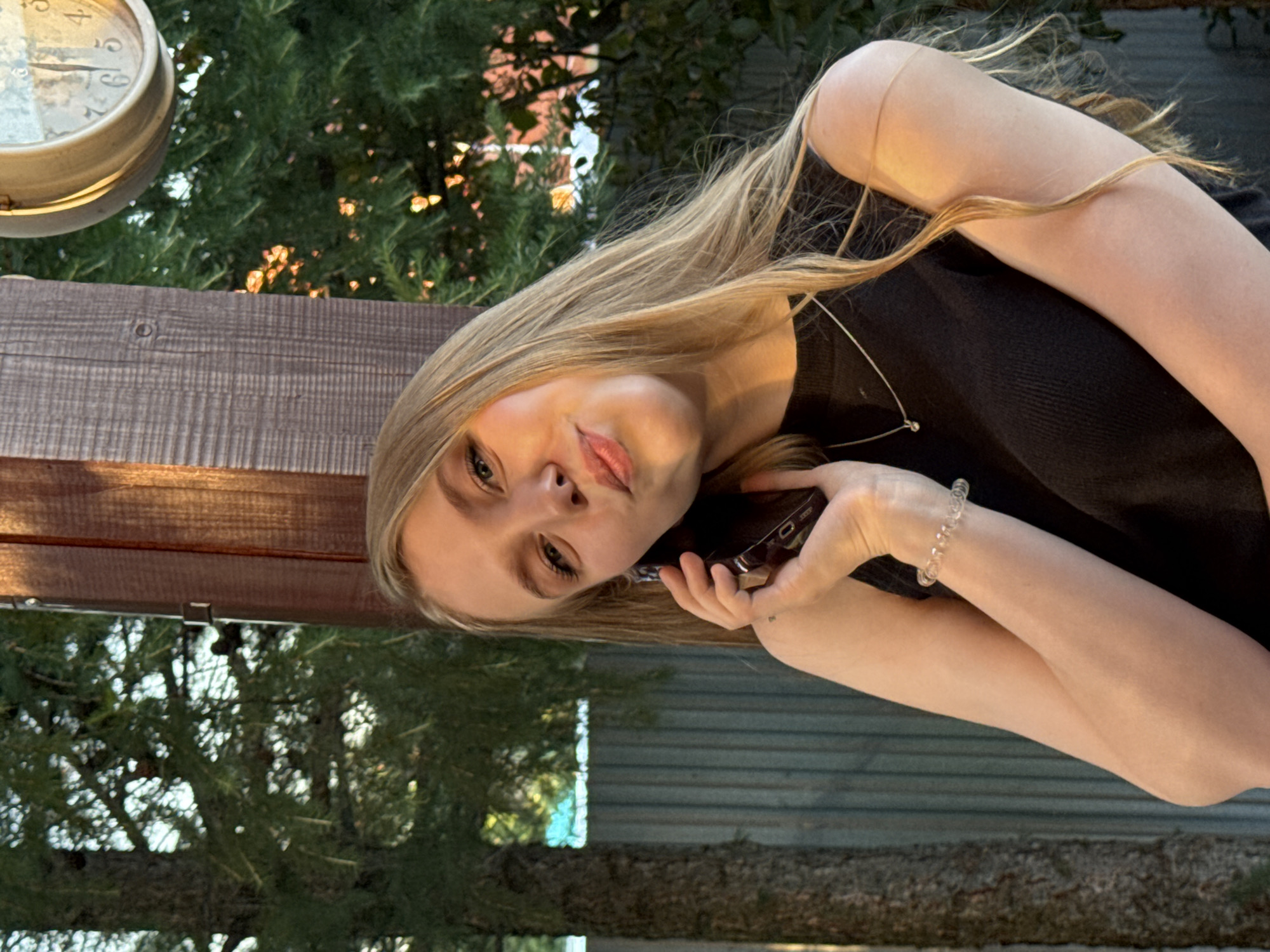 In Perm, Russia, a young Caucasian woman with long blonde hair and fair skin is captured in a medium shot, holding a dark-colored smartphone to her ear. She is wearing a sleeveless black top and a delicate silver necklace with a small pendant. Her left arm, adorned with a clear, beaded bracelet, is bent as she holds the phone. The woman is leaning against a wooden post, with a distressed grey wooden fence visible behind her. Lush green trees and foliage frame the background, with the warm glow of sunlight dappling through the leaves, suggesting it is late afternoon. A round clock with Roman numerals is partially visible in the upper right corner of the frame. The woman's expression is playful, with her lips slightly pursed as she looks towards the camera.