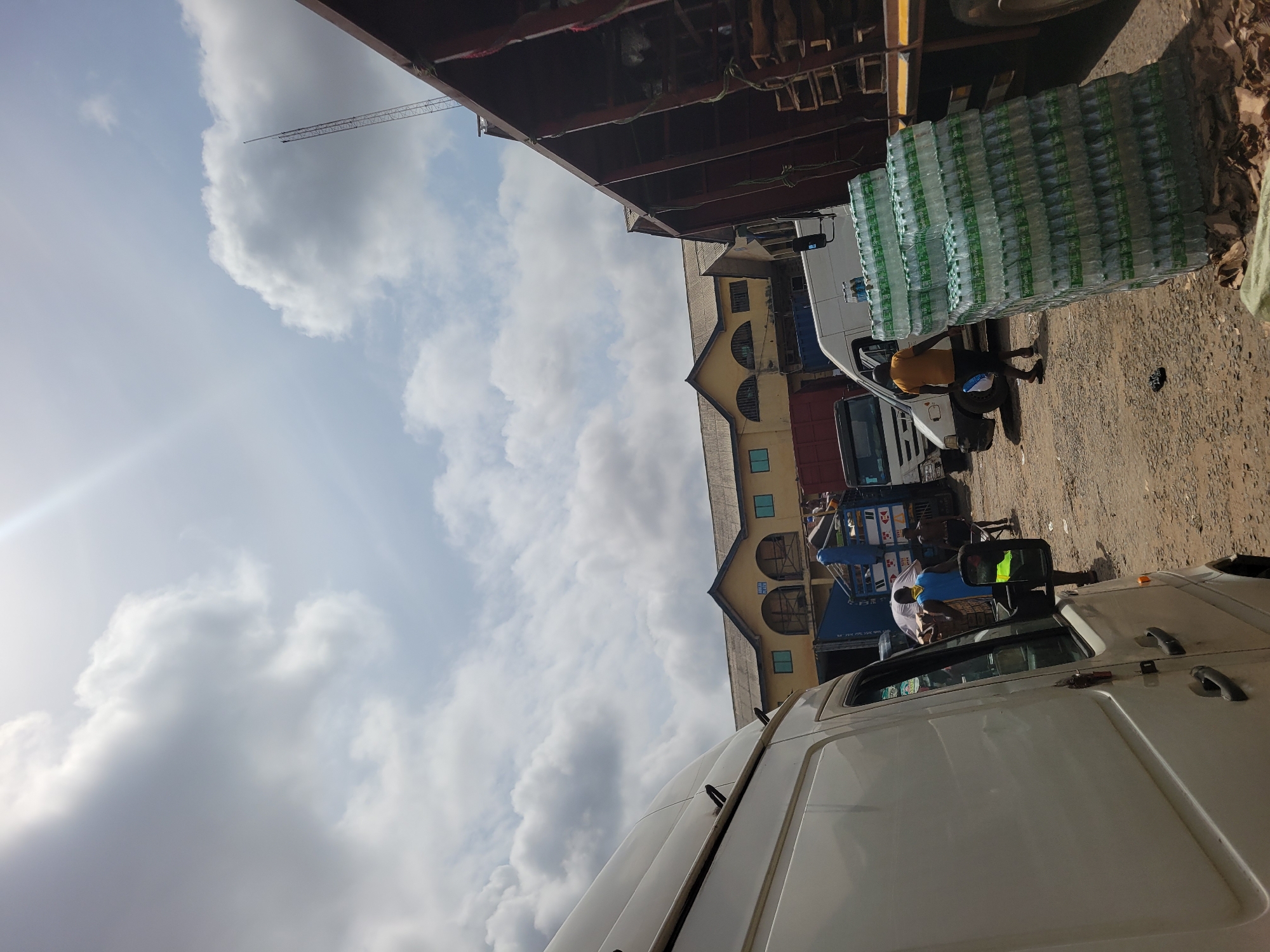 The image captures a bustling scene in what appears to be a commercial area of Onitsha, Nigeria. The sky is partially covered with white clouds. A crane can be seen in the distance. In the foreground and middle ground, there are trucks and vans parked on a dirt or gravel road. Several stacks of bottled water are visible on one of the vehicles. People are milling around the vehicles, and some seem to be loading or unloading goods. A light yellow building can be seen behind the trucks. The photo's perspective is slightly angled, with the foreground truck taking up a significant portion of the lower half of the frame.