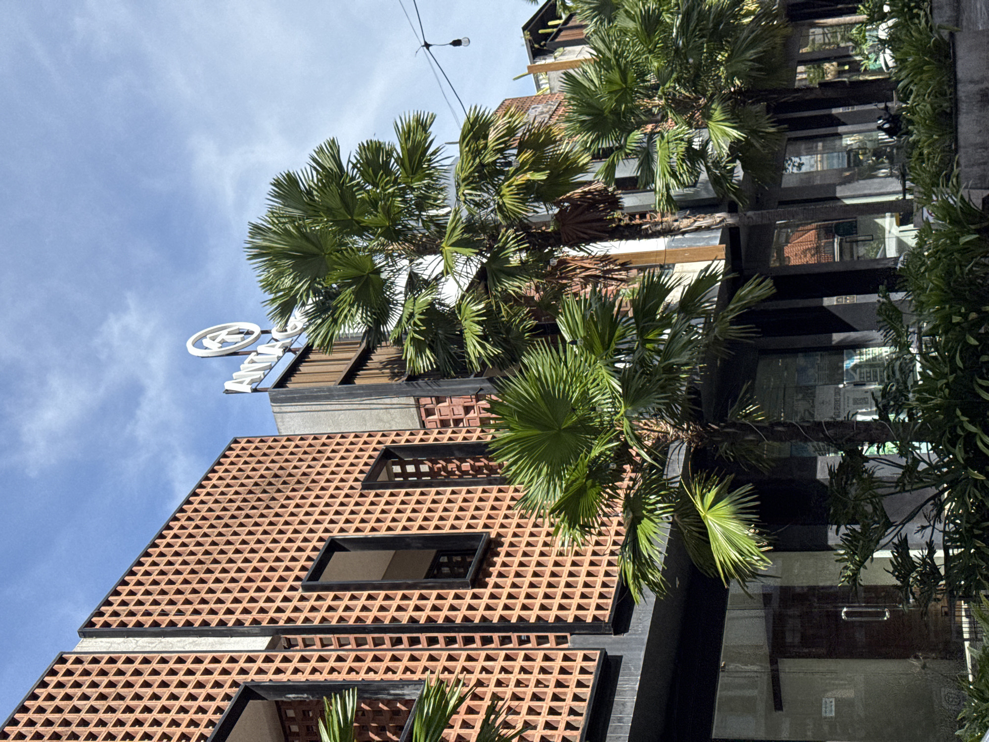 An upward, angled view showcases a building exterior in North Kuta, Indonesia. The building's facade features an open grid pattern of terracotta bricks in the bottom left, contrasted by dark window frames and a modern, glass-paneled structure to the right. Palm trees in the foreground add a tropical feel. A sign atop the building is partially visible against a blue sky.