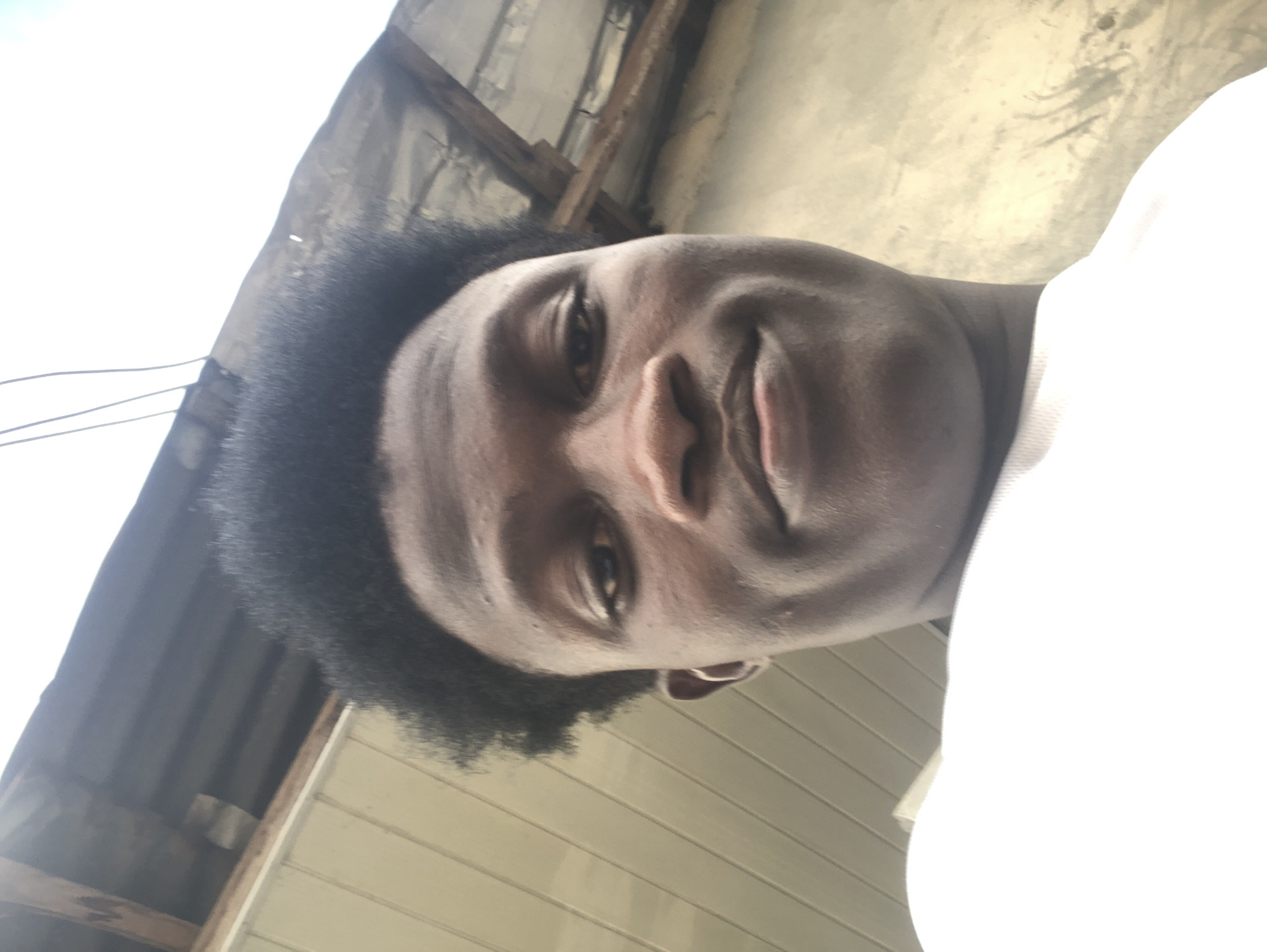 A low-angle, close-up selfie of a young Black male is captured outdoors. The subject's face, which takes up a significant portion of the frame, is turned slightly upwards, with his head tilted. He has dark, tightly curled hair, dark eyes, and a neutral expression. The background consists of a light-colored, horizontally paneled wall on the left and a weathered, grey, corrugated metal roof structure supported by wooden beams on the right. The sky is overcast and bright. The overall scene suggests an outdoor setting, possibly in Ado, Nigeria, characterized by simple, functional architecture. The image is taken from a perspective looking up at the subject.