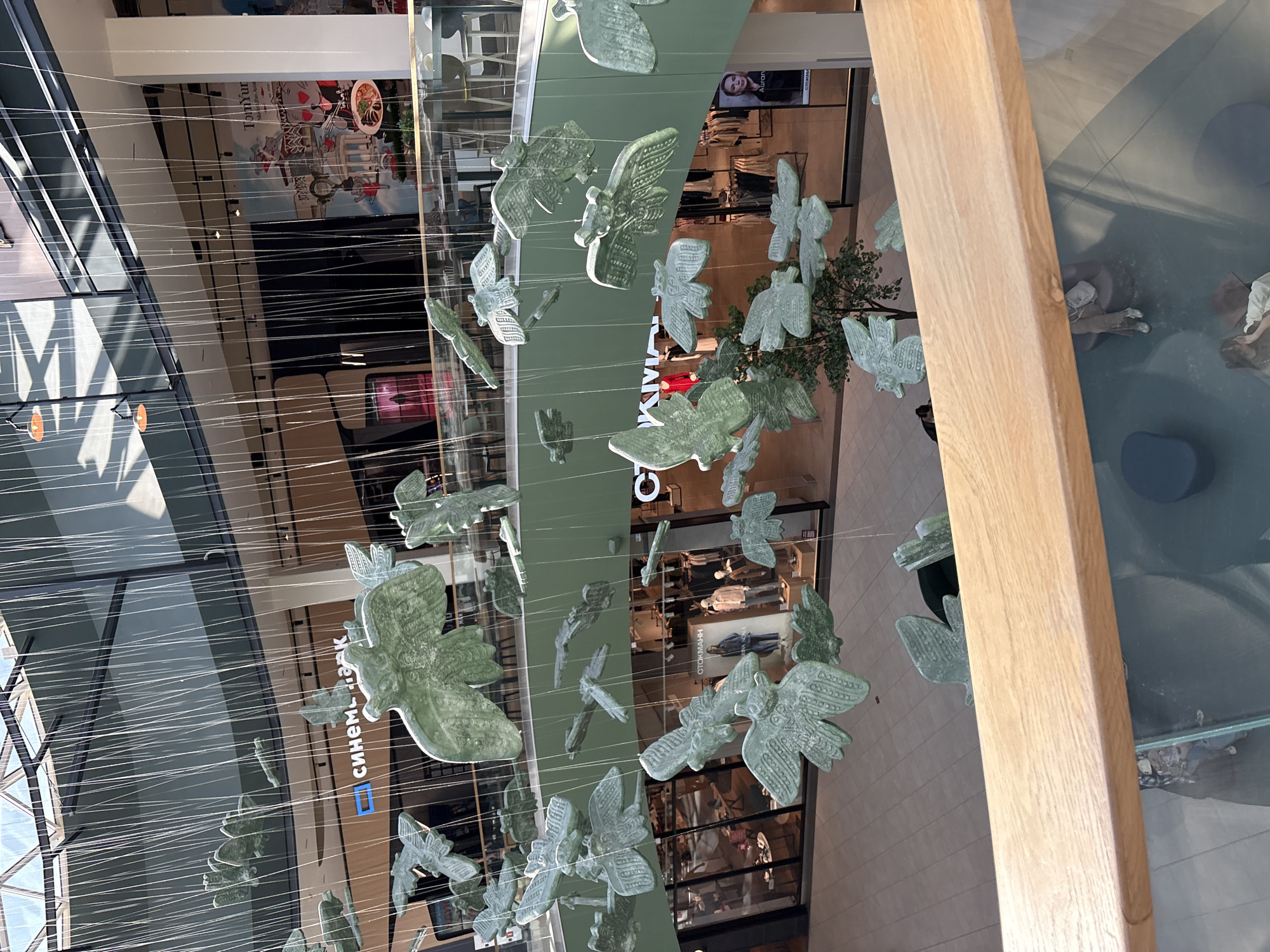 The image depicts an interior view of a multi-level shopping mall in Perm, Russia. A decorative installation of numerous stylized, light green bird-shaped objects hangs suspended from the ceiling by thin cables. Below this installation, retail store fronts are visible, including "КИНОМАКС" (KinomaX) and "СТОКМАНН" (Stockmann). Overhead, the mall's architectural features include a glass ceiling with geometric patterns. The image is taken from an elevated perspective, possibly from a balcony or upper level of the mall, with a portion of a wooden railing or beam visible in the foreground to the right. People are visible inside the stores, and the mall appears to be well-lit.