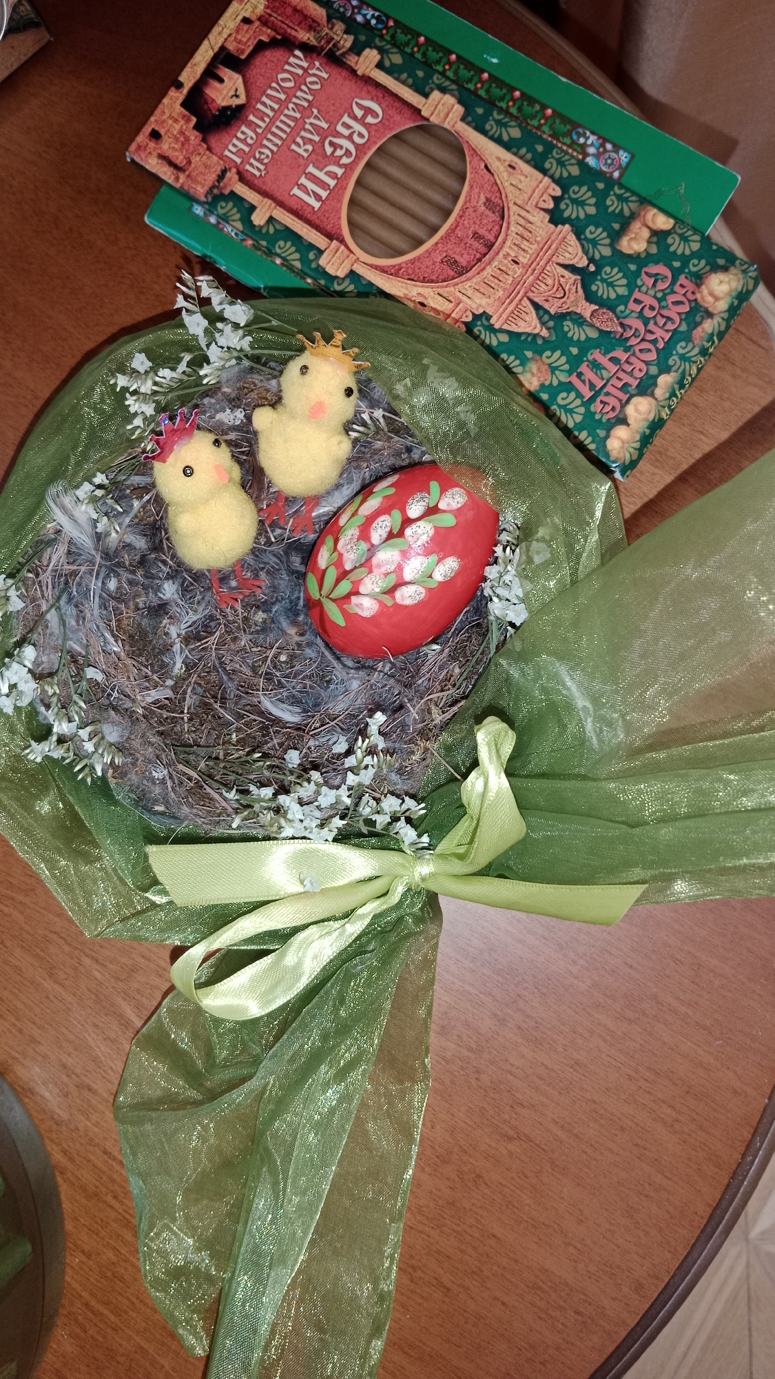 On a wooden table is a small bird's nest made of twigs and feathers, containing two small yellow chicks, each with a tiny crown, and a red Easter egg decorated with white pussy willows. The nest is wrapped in green tulle and tied with a green ribbon. Next to the nest is a box of candles labeled in Cyrillic, “Свечи для Домашней молитвы” (Candles for Home Prayer) and “Воскресные свечи” (Sunday candles). The presence of Easter decorations and candles suggests a religious theme, possibly related to Russian Orthodox traditions. The location cannot be determined from the image.