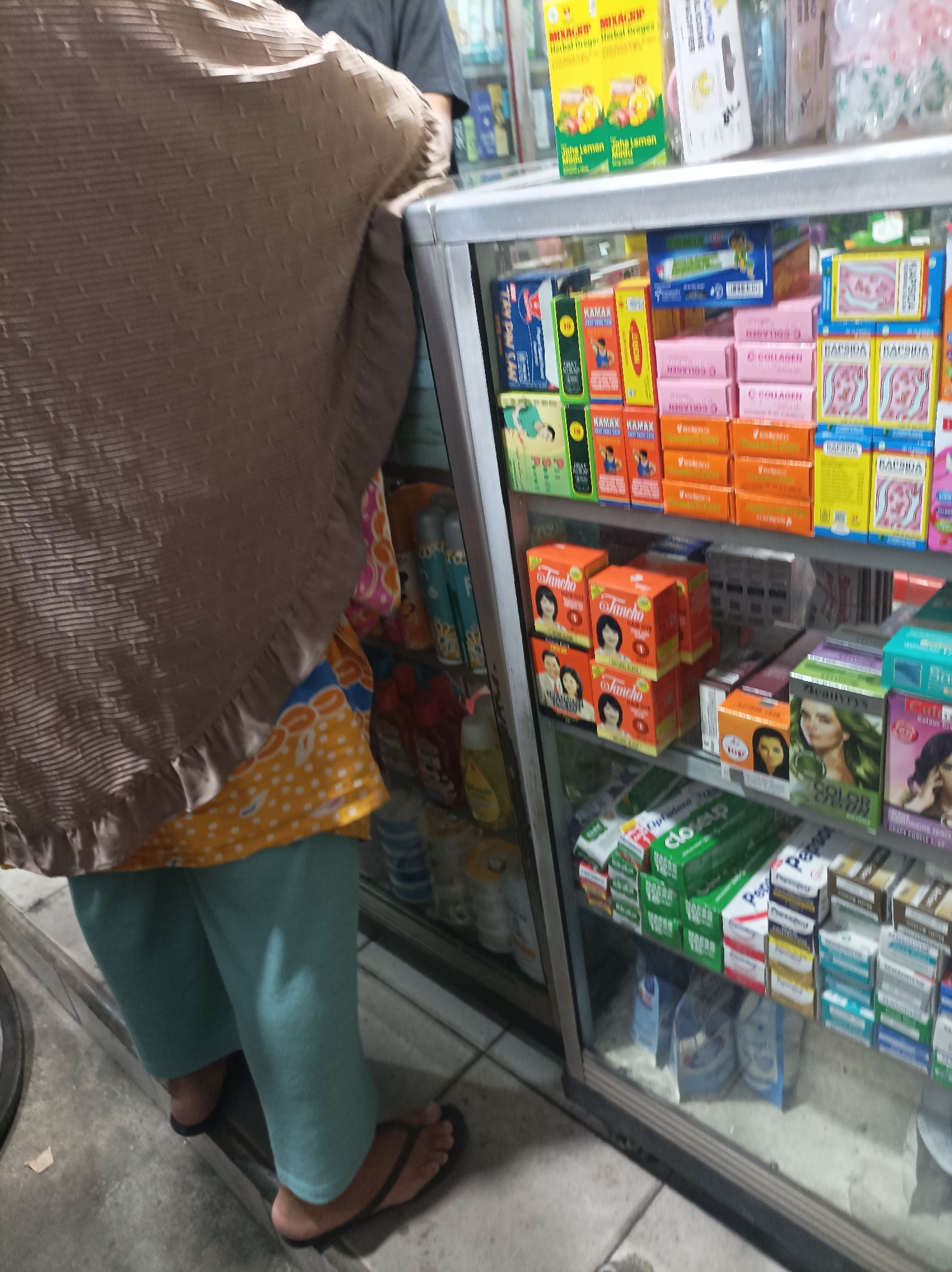 A person wearing a brown hijab, an orange and yellow patterned top, and light blue pants stands in front of a glass-enclosed display shelf filled with various health and beauty products. The display shelf appears to be in a small shop or pharmacy. The person's feet are visible, wearing dark flip-flops. The shelves are organized with boxed items, including medications, skincare products, and personal care items. The products are in colorful packaging with text in what appears to be Indonesian. The lighting suggests an indoor setting, possibly during the day or under artificial lighting. The background shows more shelves and products, and a glimpse of what might be a street outside the shop. The image is taken from a slightly elevated angle, looking down at the person and the display. The specific location is a shop in the city of West Jakarta, Indonesia.