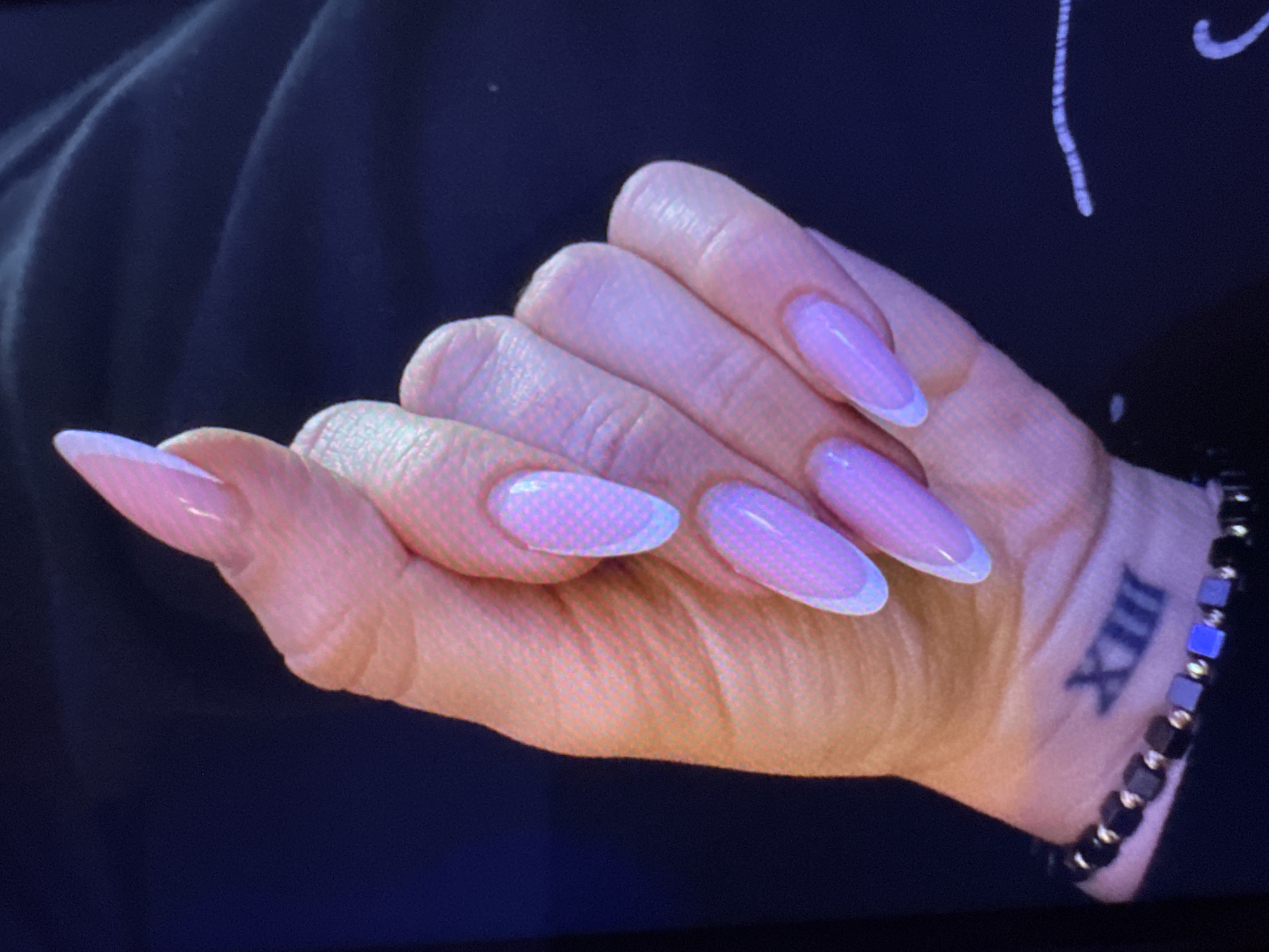 The image displays a close-up of a person's hand with artificial nails. The nails are long and almond-shaped, with a French manicure design featuring a sheer pink base and white tips. Some of the nails have a subtle, light purple pattern with a dotted texture. On the wrist, there is a black beaded bracelet with small square and round beads, and a tattoo of Roman numerals "XIII". The background is dark and indistinct, with a hint of fabric texture. There is no discernible information about the location in Plovdiv, Bulgaria, as the image is a close-up of a hand and the background lacks any identifying features of a specific place.