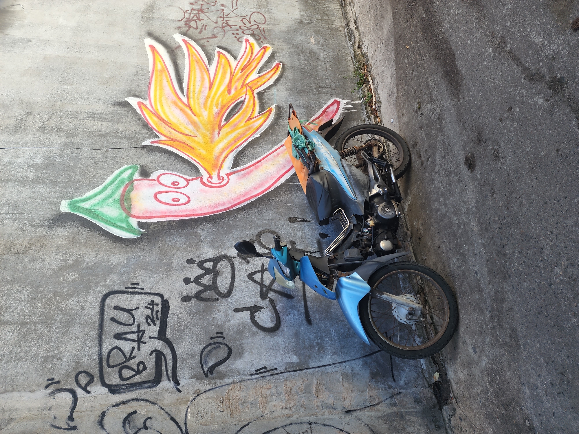 A light blue motorcycle is parked on a paved street in front of a concrete wall covered in graffiti. The graffiti includes a cartoonish drawing of a chili pepper with flames coming out of its mouth, as well as various words and symbols. The motorcycle is a standard commuter model with a black seat and a slightly worn appearance. The street appears to be in an urban or semi-urban setting. The image was taken on a sunny day, with shadows visible on the ground. The location is identified as Phu Thuy Ward, Vietnam.