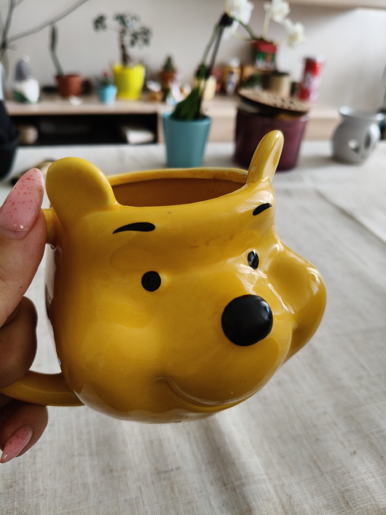 A yellow Winnie the Pooh mug is held in a person's hand with a light pink manicure. The mug is ceramic and features the character's face with black features. The background shows a blurry assortment of houseplants in various pots, a lidded jar, and other decorative objects on a light-colored surface. The scene takes place indoors. There is no information available about the location in the city of Plovdiv, Bulgaria.