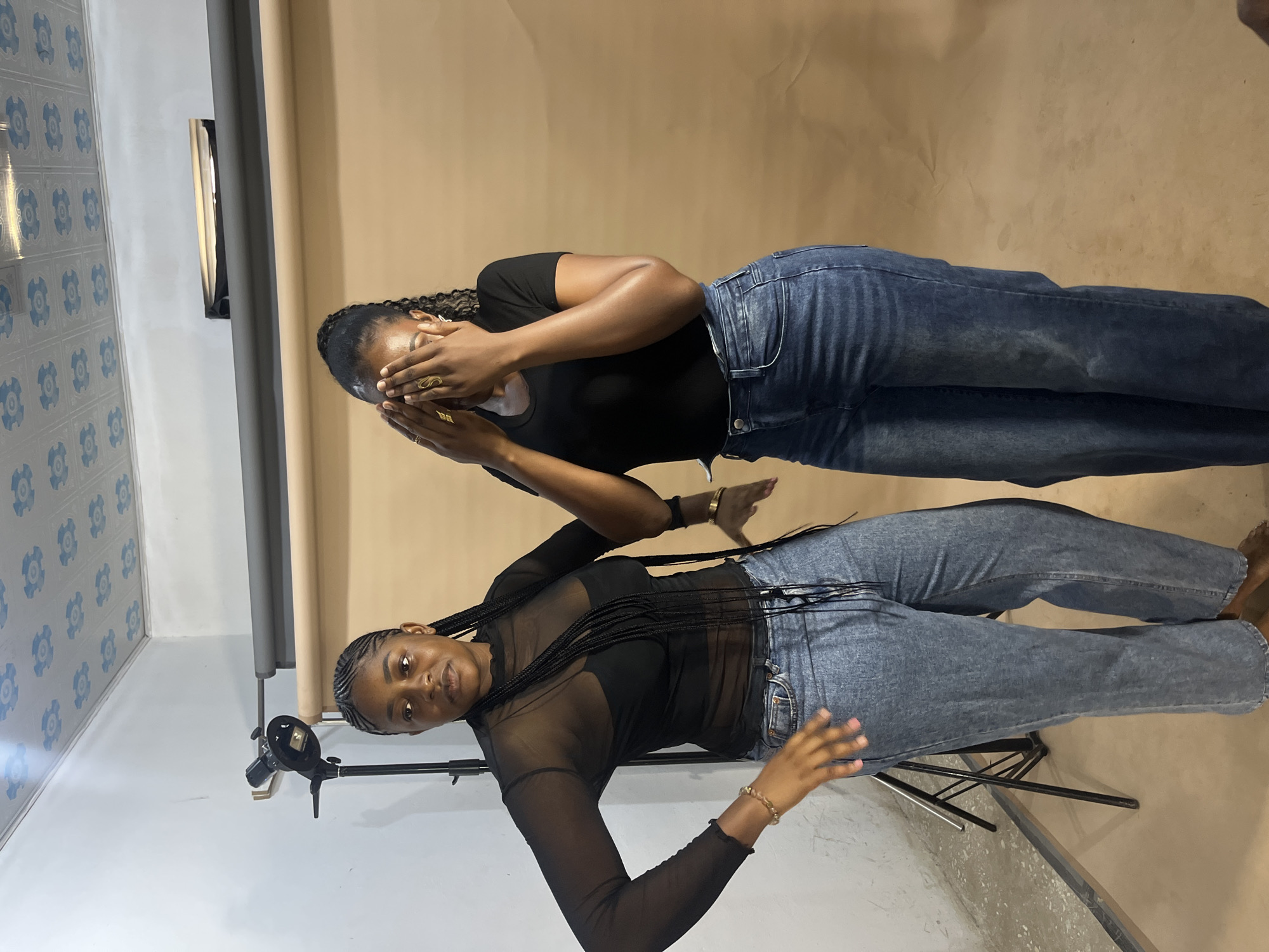 In Gura Top, Nigeria, within a studio setting featuring a plain, tan backdrop and a white wall, two women are photographed. The woman on the left, with braided hair, wears a sheer black long-sleeved top over a black bra and blue denim jeans. She stands with her left hand on her hip and her right hand extended forward, palm open. The woman on the right, also with braided hair, is dressed in a black t-shirt and blue denim jeans. She covers her face with both hands. A lighting rig is visible in the background to the left of the woman on the left. The ceiling is decorated with a repeating blue floral pattern.