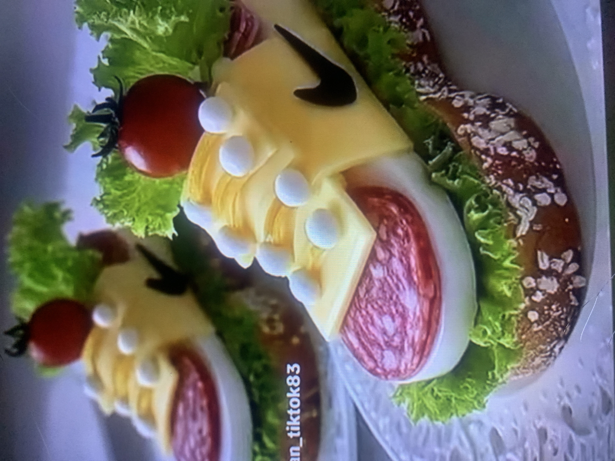 The image shows two food items shaped like sneakers, placed on plates. Each sneaker-shaped food item consists of a slice of bread as the sole, topped with lettuce, a slice of hard-boiled egg, a slice of salami, and then layered with yellow cheese to form the upper part of the shoe. Small white balls, likely mozzarella balls or small pieces of cheese, are placed on the cheese layer to represent laces. A cherry tomato and lettuce are used as decorative elements at the back of each shoe. A black Nike logo is visible on the side of the cheese "shoe" on the right. The background is blurred and appears to be an indoor setting. Text overlay on the left side of the image reads "an_tiktok83". There is no information in the image to suggest a location in Almaty, Kazakhstan.