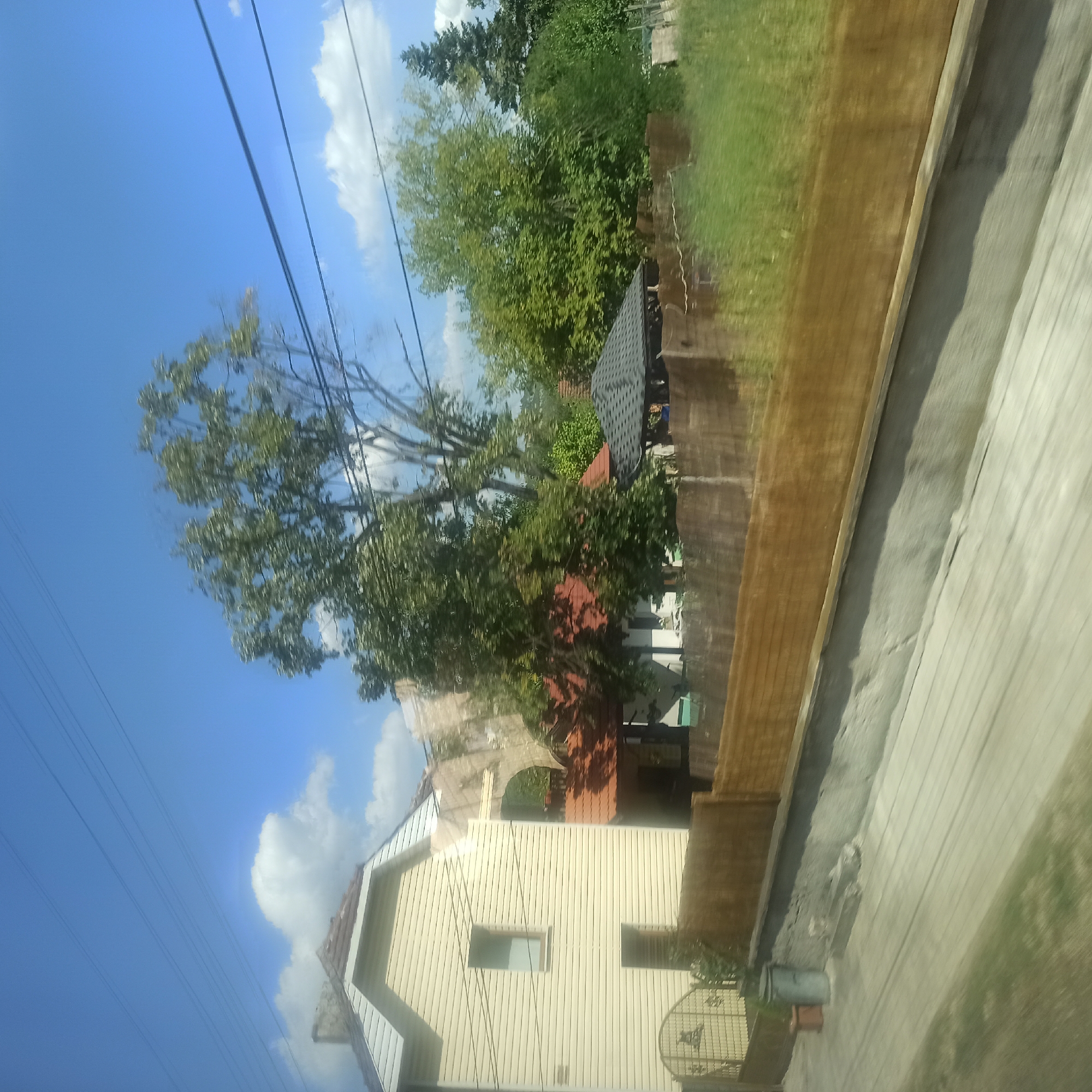 The image shows a residential area on a sunny day, with a clear blue sky and scattered clouds. In the foreground, a light-colored, horizontal wooden fence runs across the frame. Behind the fence, a light yellow, two-story house with white trim and a brown roof is visible on the left. The house has a single window on the upper floor and a dark doorway on the ground floor. To the right of the house, a covered outdoor seating area with a dark grey, multi-faceted roof is present. Trees with green foliage are abundant throughout the scene, with a large tree prominently positioned behind the house and seating area. There is a grey trash can and a small reddish pot near the fence. The ground in front of the fence appears to be a paved path with a light grey, textured surface. The background shows more trees and vegetation, suggesting a suburban or rural setting. The image appears to have been taken from a moving vehicle, as indicated by the slight motion blur. The scene is located in Zlatna Panega, Bulgaria.